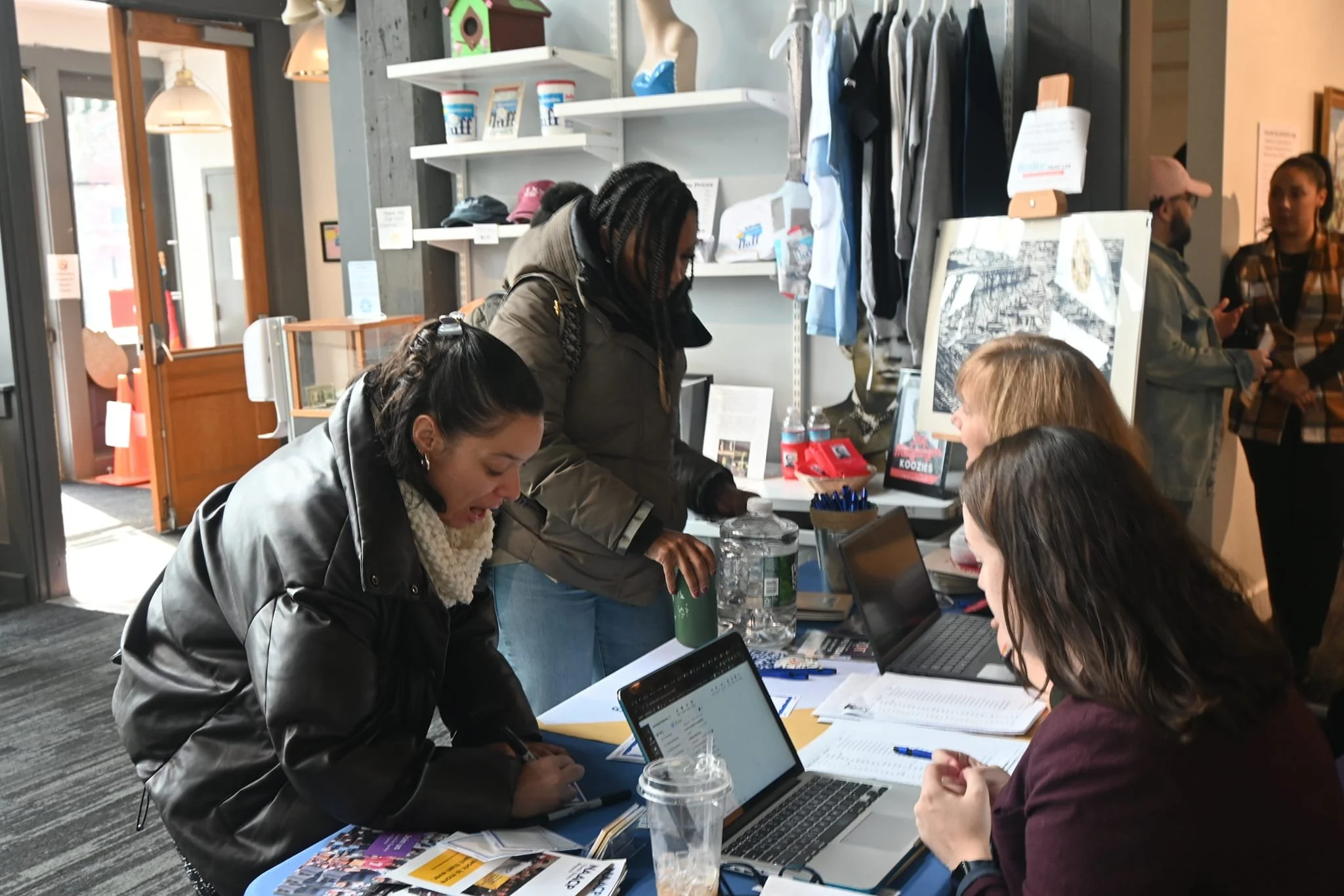 Women working at a registration desk inside a public event at a local museum. The women are focused on laptops and paperwork, greeting guests, and have pamphlets and artwork displayed around them.