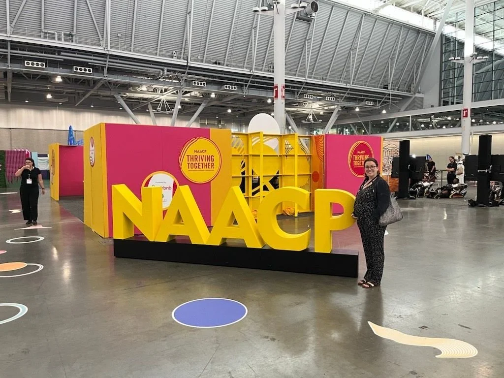 A woman standing next to large yellow 3D letters spelling NAACP at an indoor event with a colorful background and people in the distance.