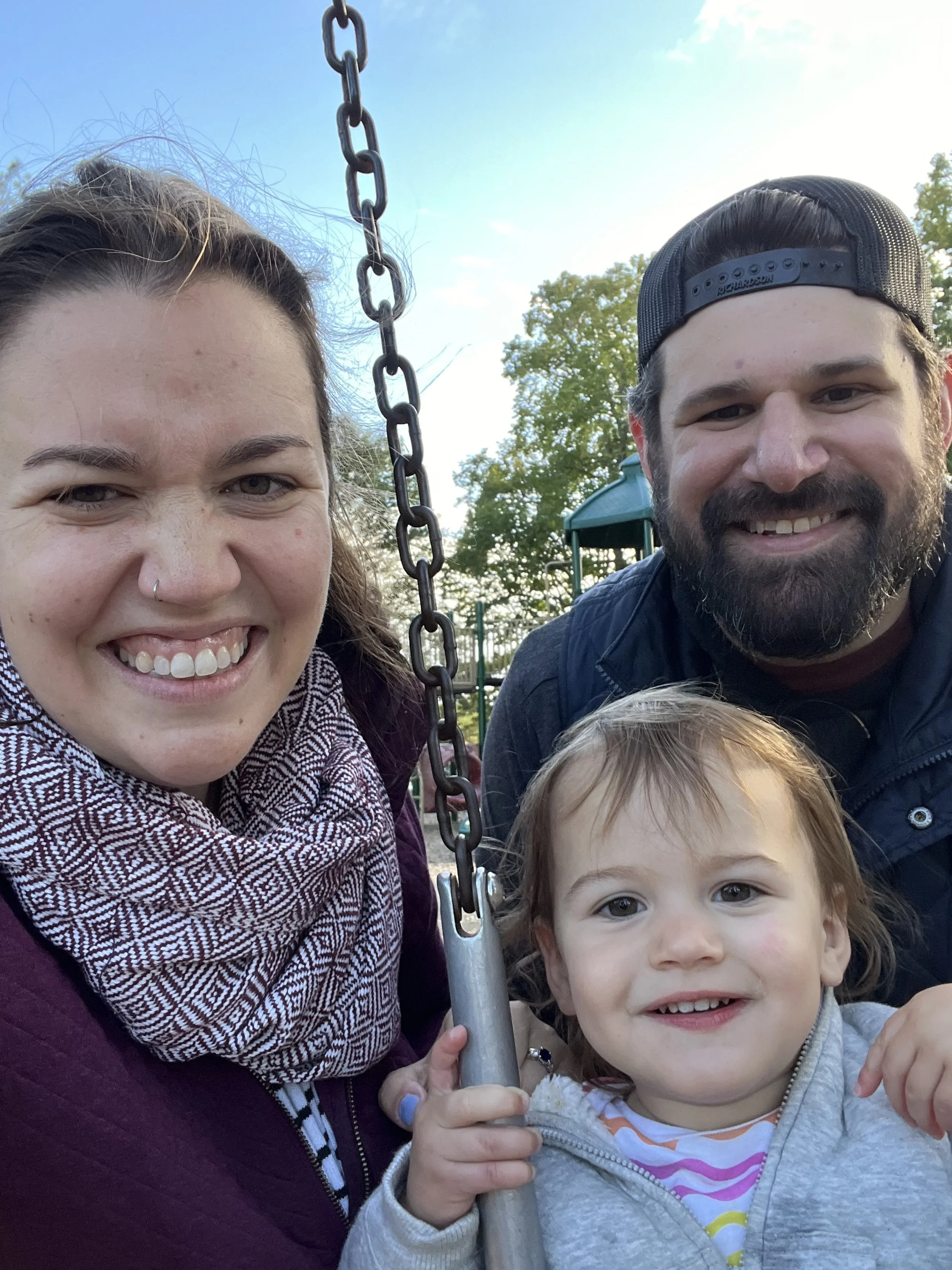 A family of three smiling at the camera on a swing at a park, with trees and playground equipment in the background.