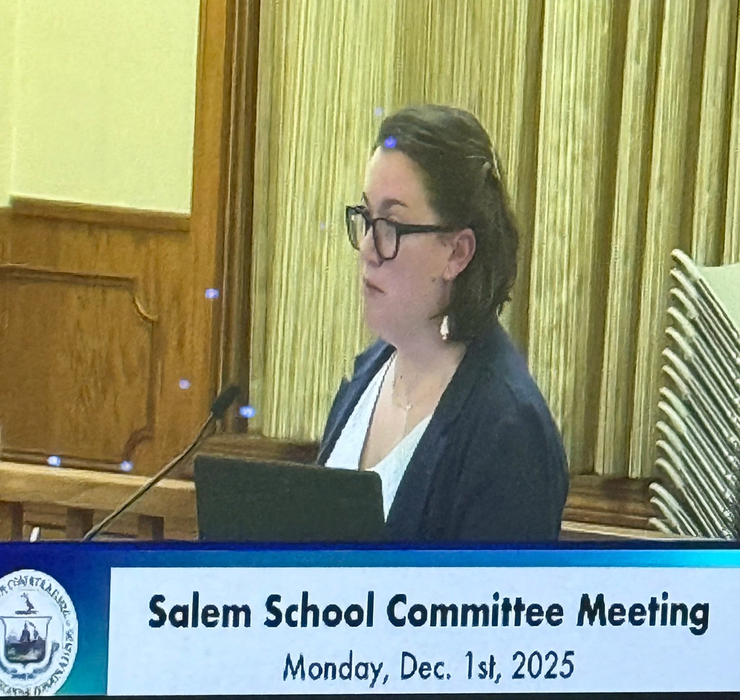 A woman with glasses and dark hair speaking at a Salem School Committee Meeting on December 1st, 2025.