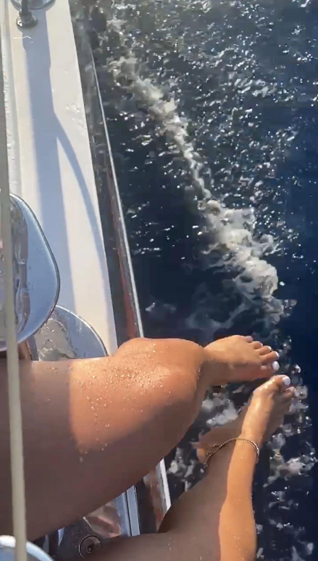 Person relaxing on a boat, with legs extended, sitting near the edge, with water and a trail of bubbles behind.