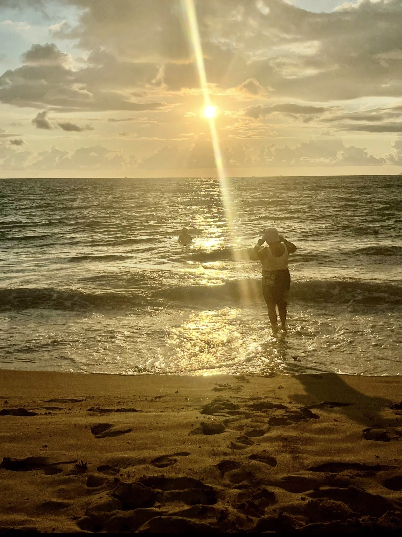 Sunset at the beach with three people in the water, one of whom is taking a photo, and footprints on the sandy shore.