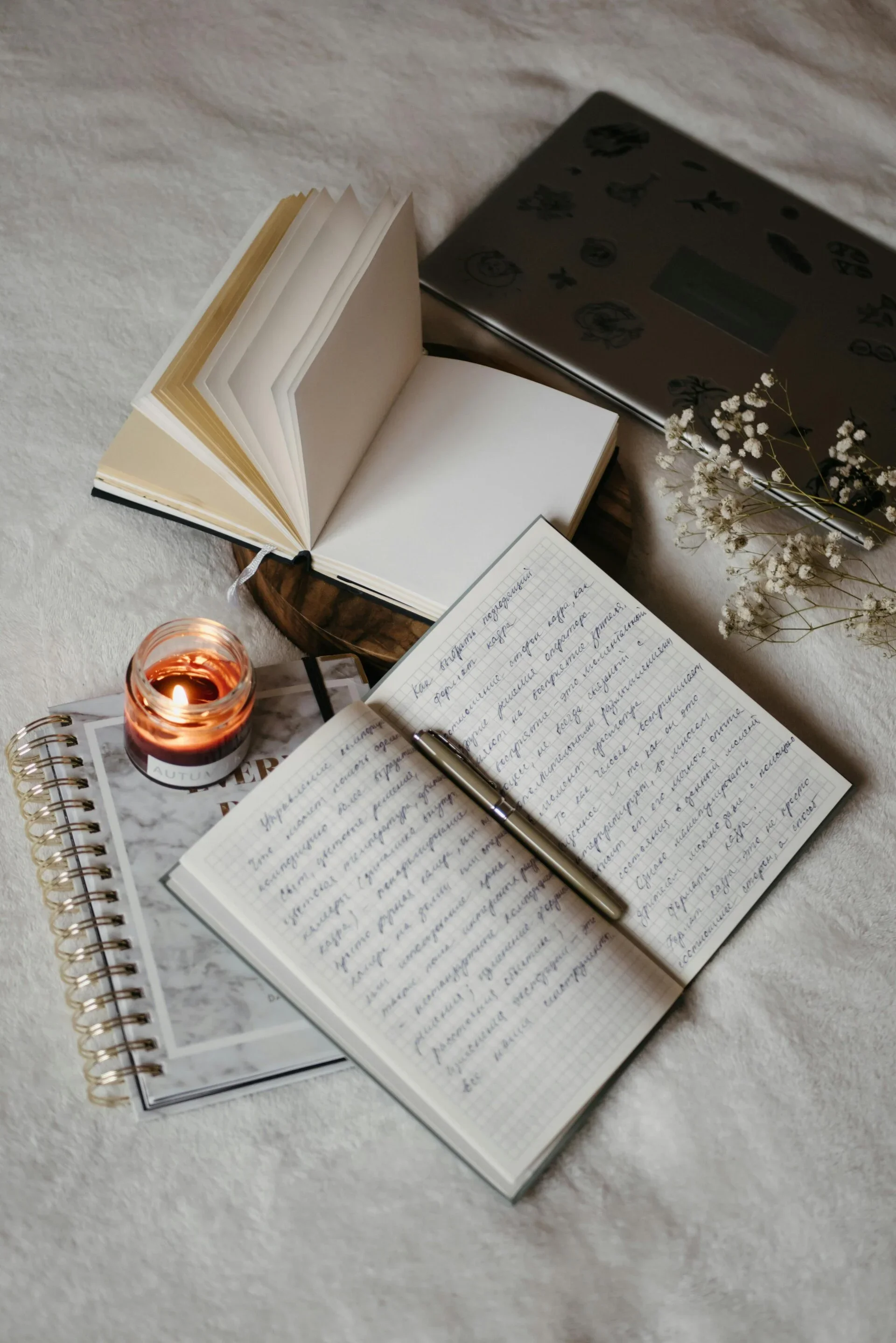 Open notebooks, a candle, a pen, and dried flowers arranged on a white textured surface.