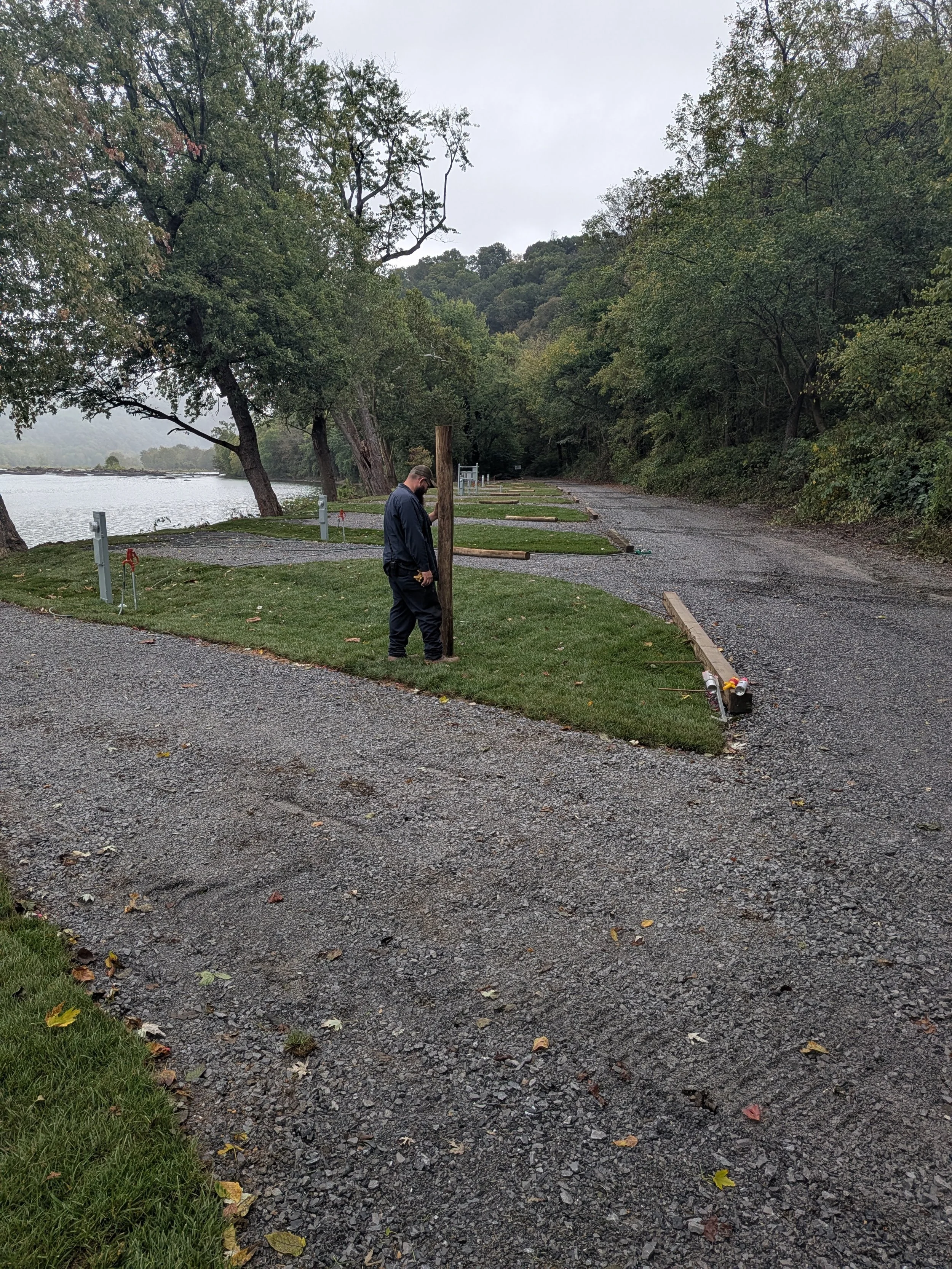 A man planting a wooden pole along a gravel path near a body of water, with several similar poles and frames to be installed, surrounded by trees on a cloudy day.