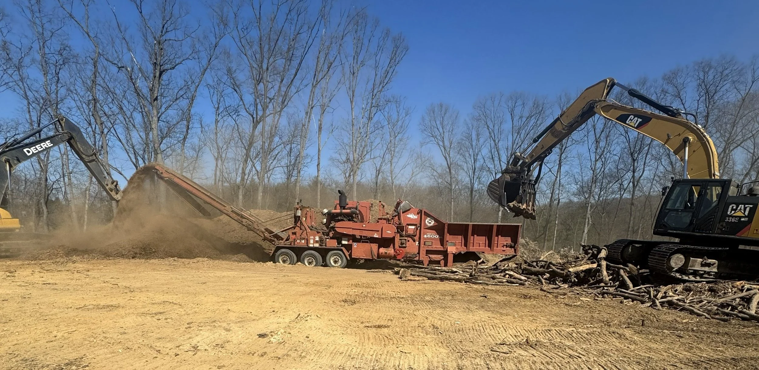 Construction site with excavators and a wood chipper processing logs against a background of leafless trees and a clear blue sky.