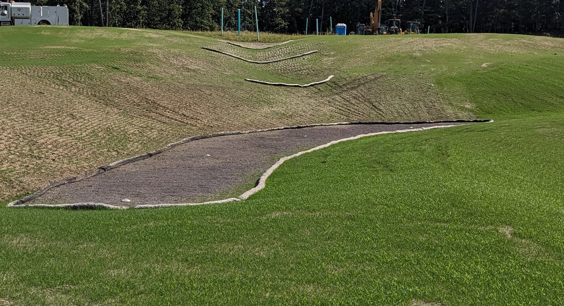 A miniature golf course with a green and a brown, sandy putting area with stone border, in a grassy outdoor setting with trees and construction equipment in the background.