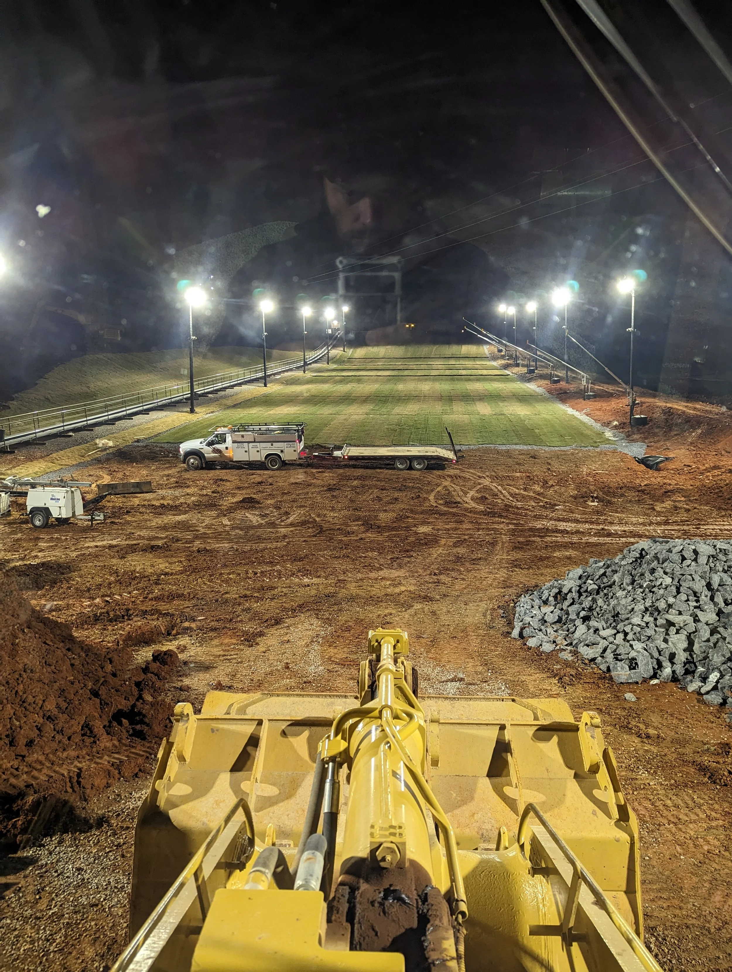 Nighttime view from a construction vehicle looking at a sports field with bright lights. Construction equipment and gravel are visible in the foreground.