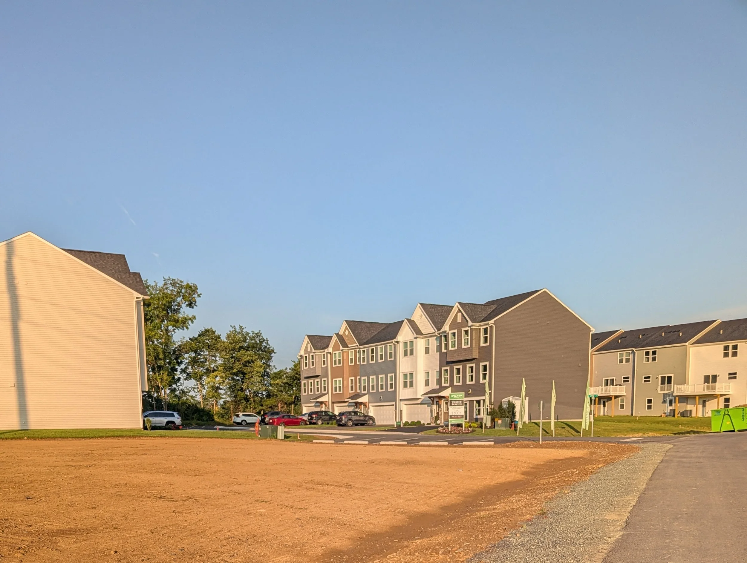 A row of modern three-story townhouse buildings with parking lot and cars in front, under a clear blue sky.