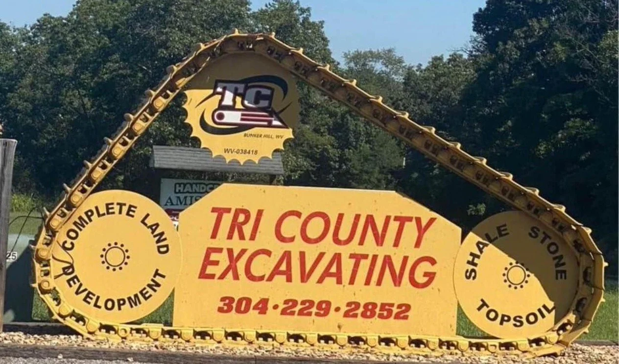 Sign for Tri County Excavating with a large yellow and black excavator arm design featuring the company name, phone number, and services like complete land development, shale, stone, and topsoil.