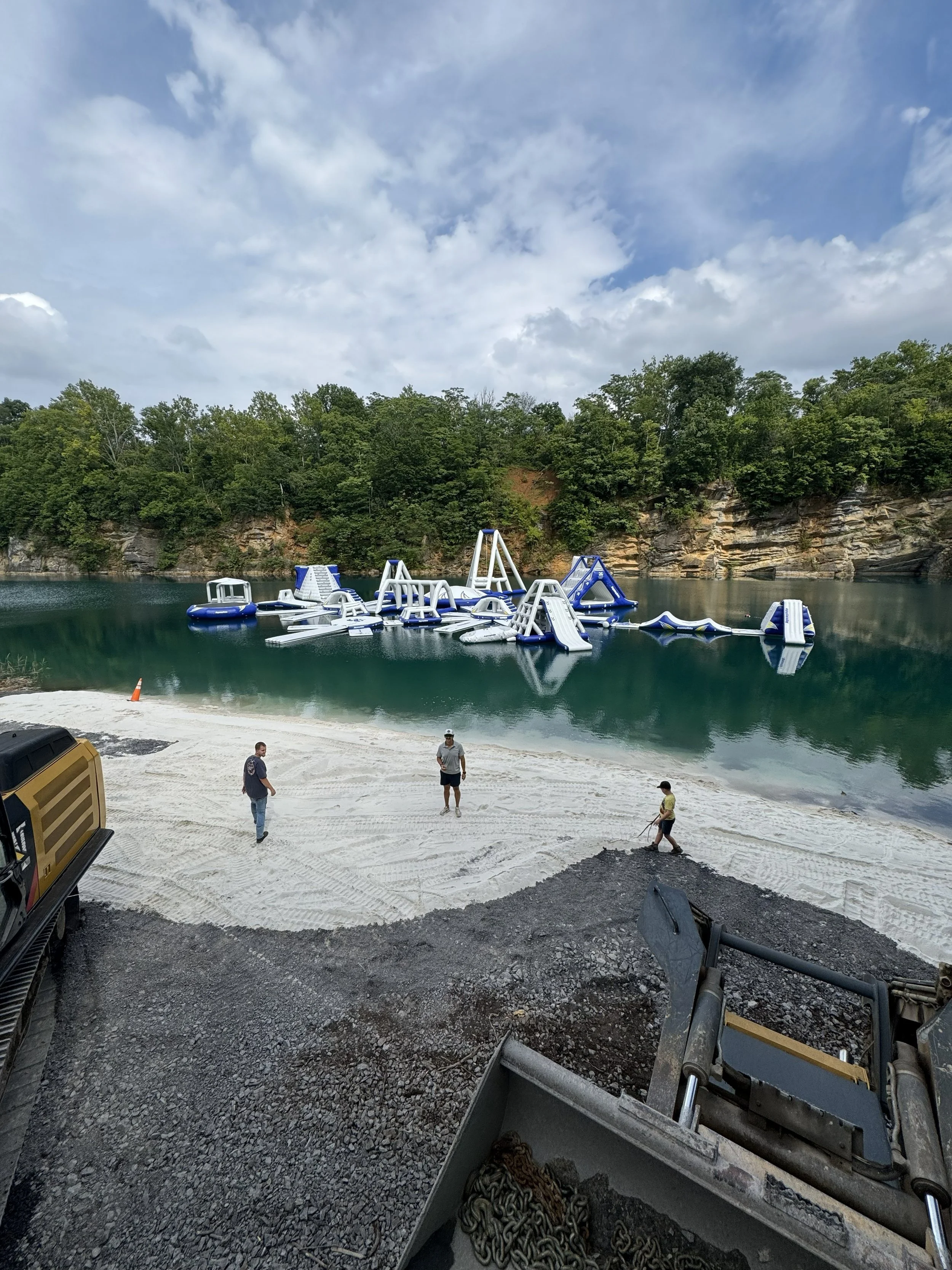 View of an outdoor waterpark with inflatable water slides and obstacles set up in a body of water, surrounded by green trees and rocky hills, with three people standing on a sandy and gravel area near construction equipment.