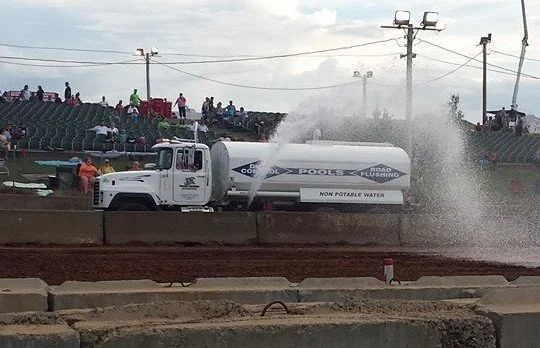 A water truck spraying water on a dirt track at a stadium with spectators and power poles in the background.