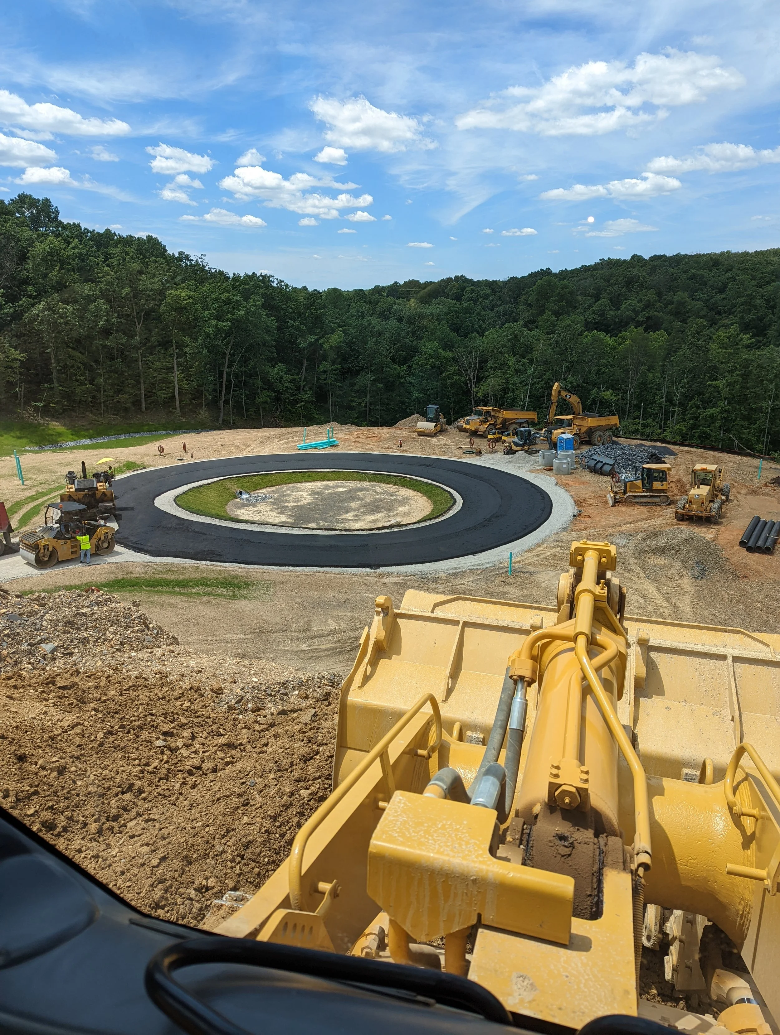View from a construction vehicle looking over a circular road under construction, with construction equipment and workers, surrounded by green trees and blue sky with clouds.