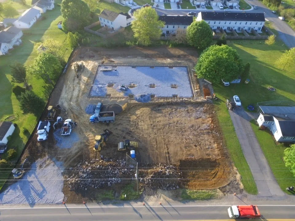 Construction site in a residential area with a newly poured concrete foundation, construction equipment, and parked vehicles surrounding the site.