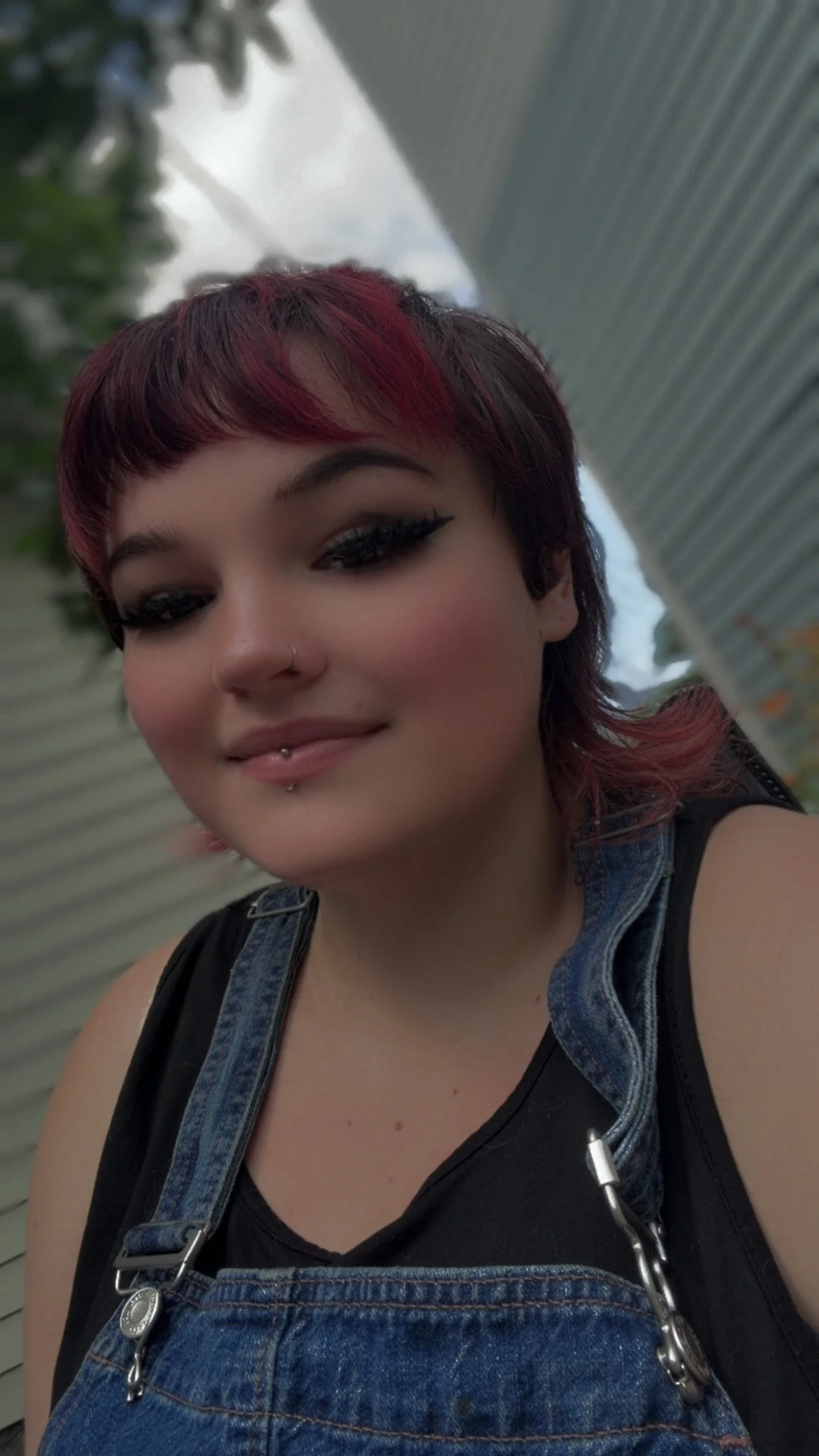 A white person with short red hair wearing a black tank top and denim overalls.