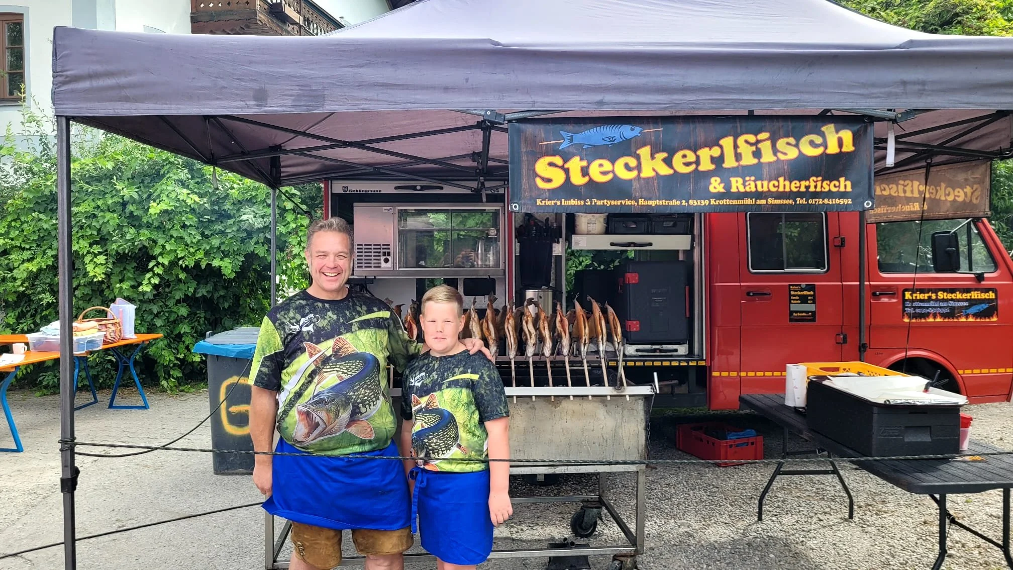 Zwei Männer stehen vor einem Verkaufswagen, der geräucherte Fisch verkauft, mit einem Schild mit der Aufschrift 'Steckerfisch & Räucherfisch'.