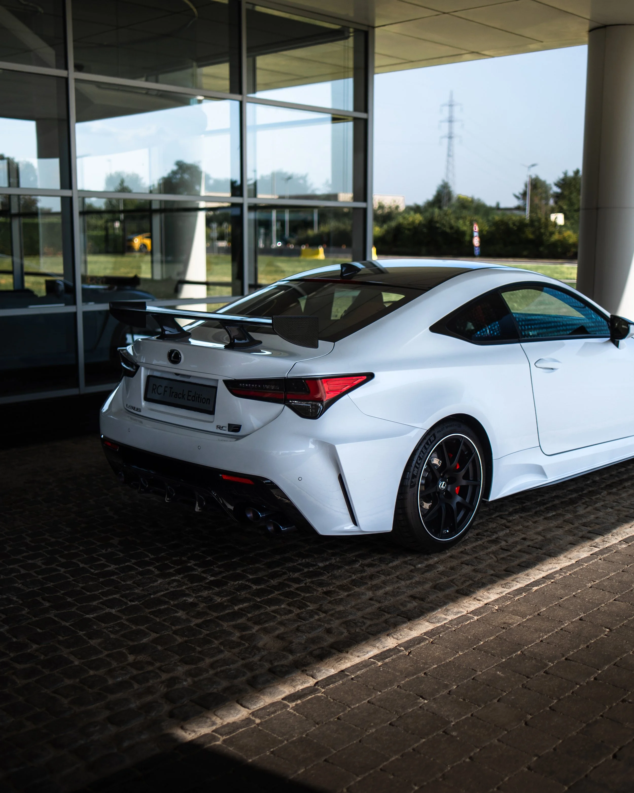White Lexus RC F sports car with black accents and a large rear wing, parked outside a modern glass building.