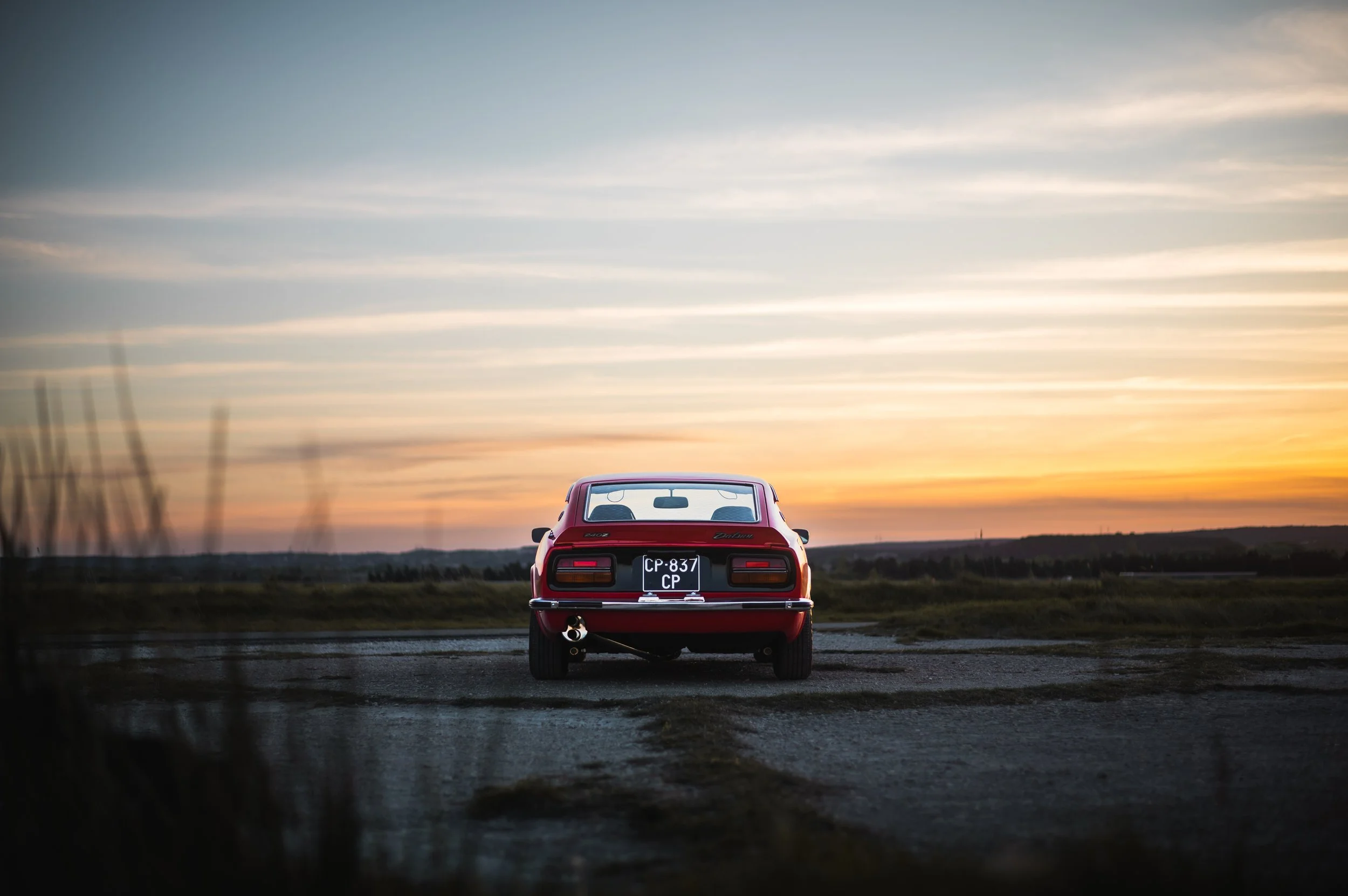 Red Datsun 240Z parked on a rural road at sunset with a colorful sky and open landscape in the background.
