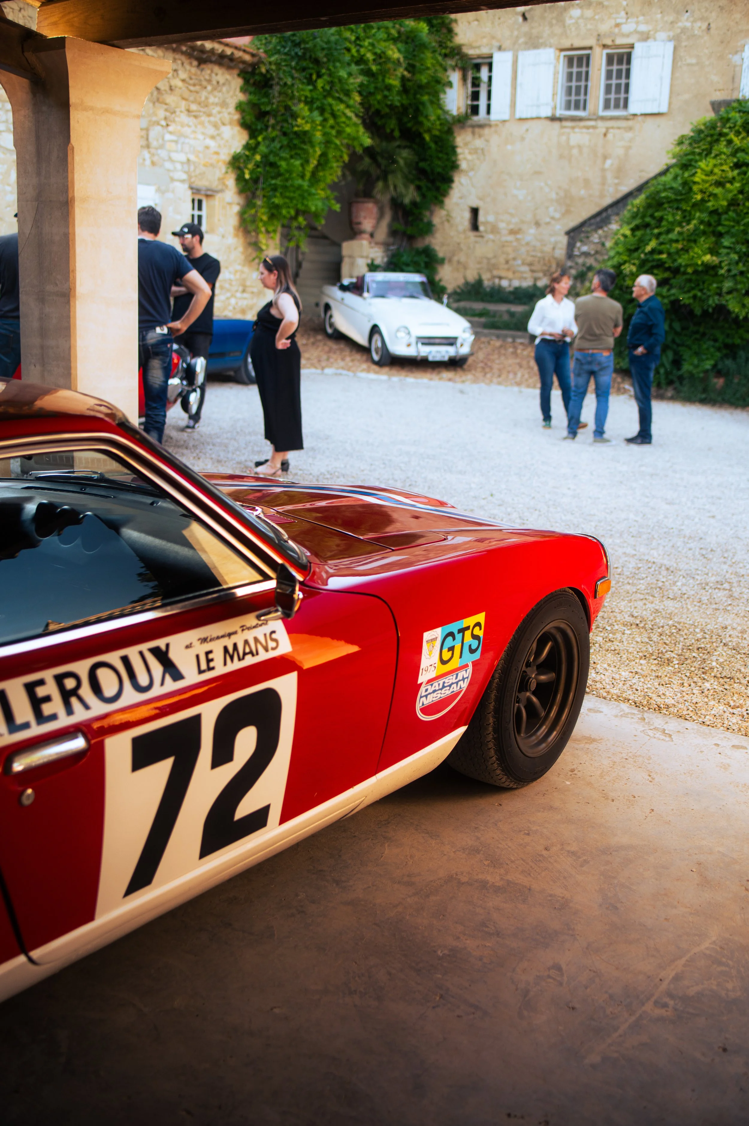 A classic red race car with the number 72 on its side is parked in a garage. In the background, several people are standing outside on a gravel driveway, engaged in conversations, with an antique white car and a blue car parked nearby. The setting ap