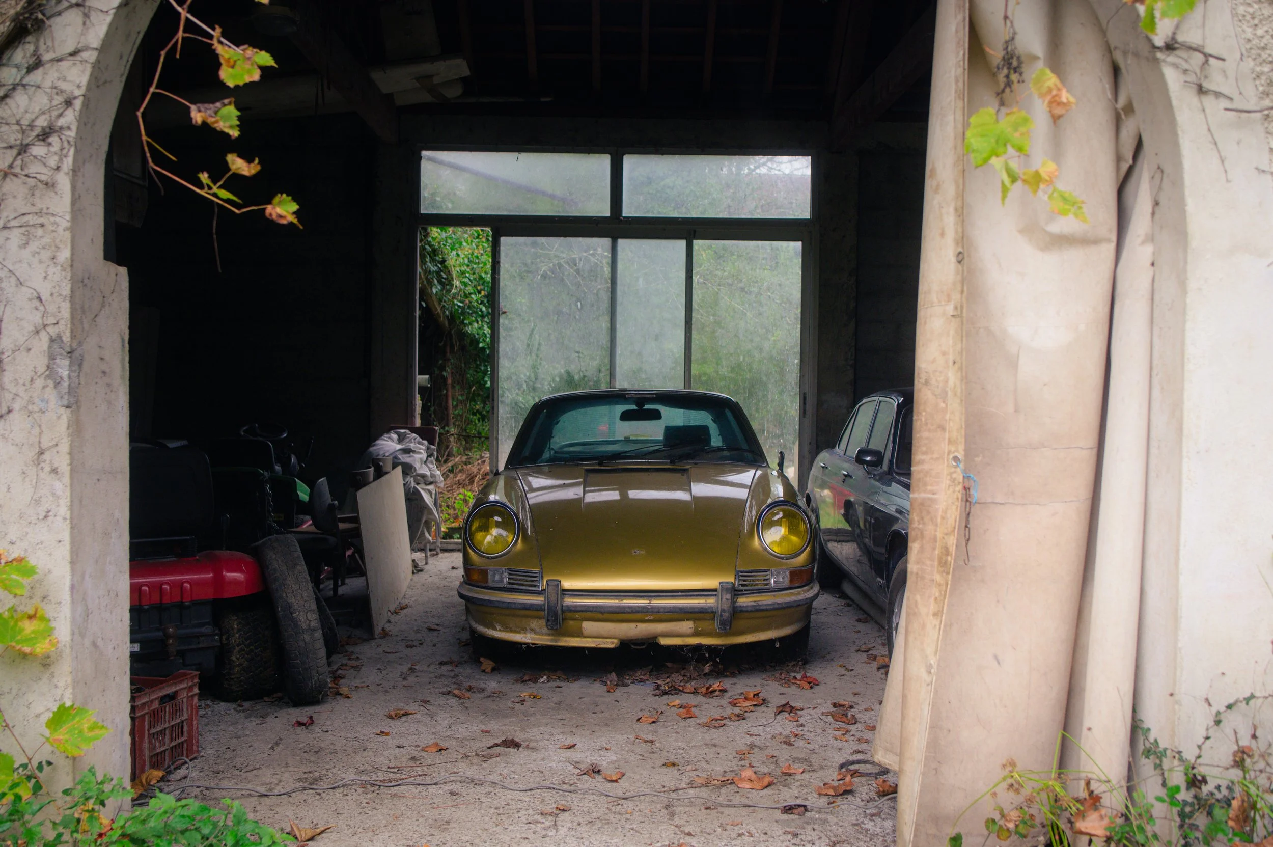 A vintage 1972 Porsche 911 2.4T Oil Trap parked inside a rustic garage with a roll-up door and a large window, surrounded by clutter including a red lawnmower, tires, and miscellaneous items.