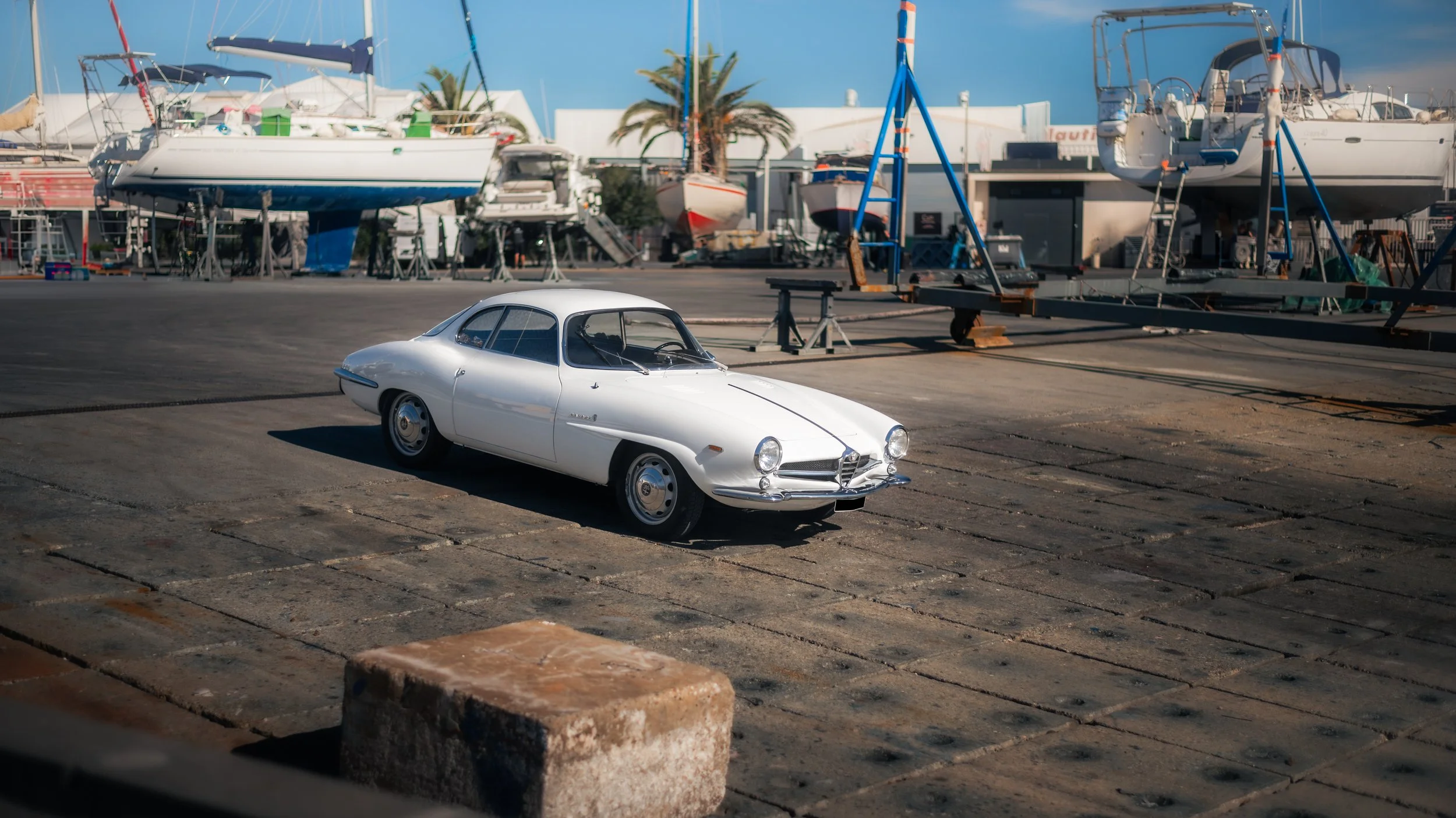 A white Alfa Romeo Giulia Sprint Speciale parked on a dry dock or marina area with boats and sailboats on stands in the background, palm trees, and a partly cloudy sky.