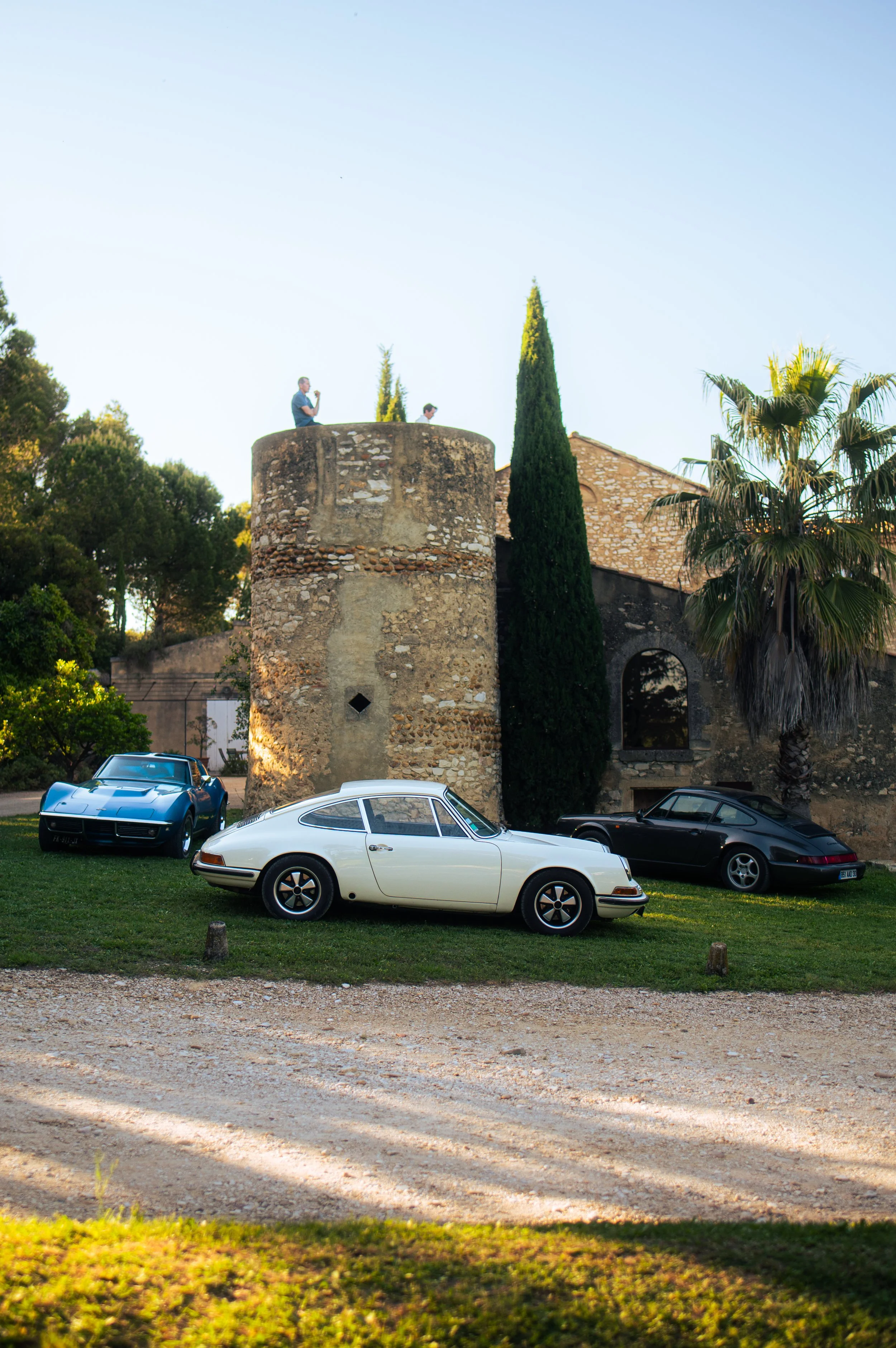 Three classic cars parked on grass in front of an old stone building with a tower, surrounded by tall trees, with two people standing on top of the tower.