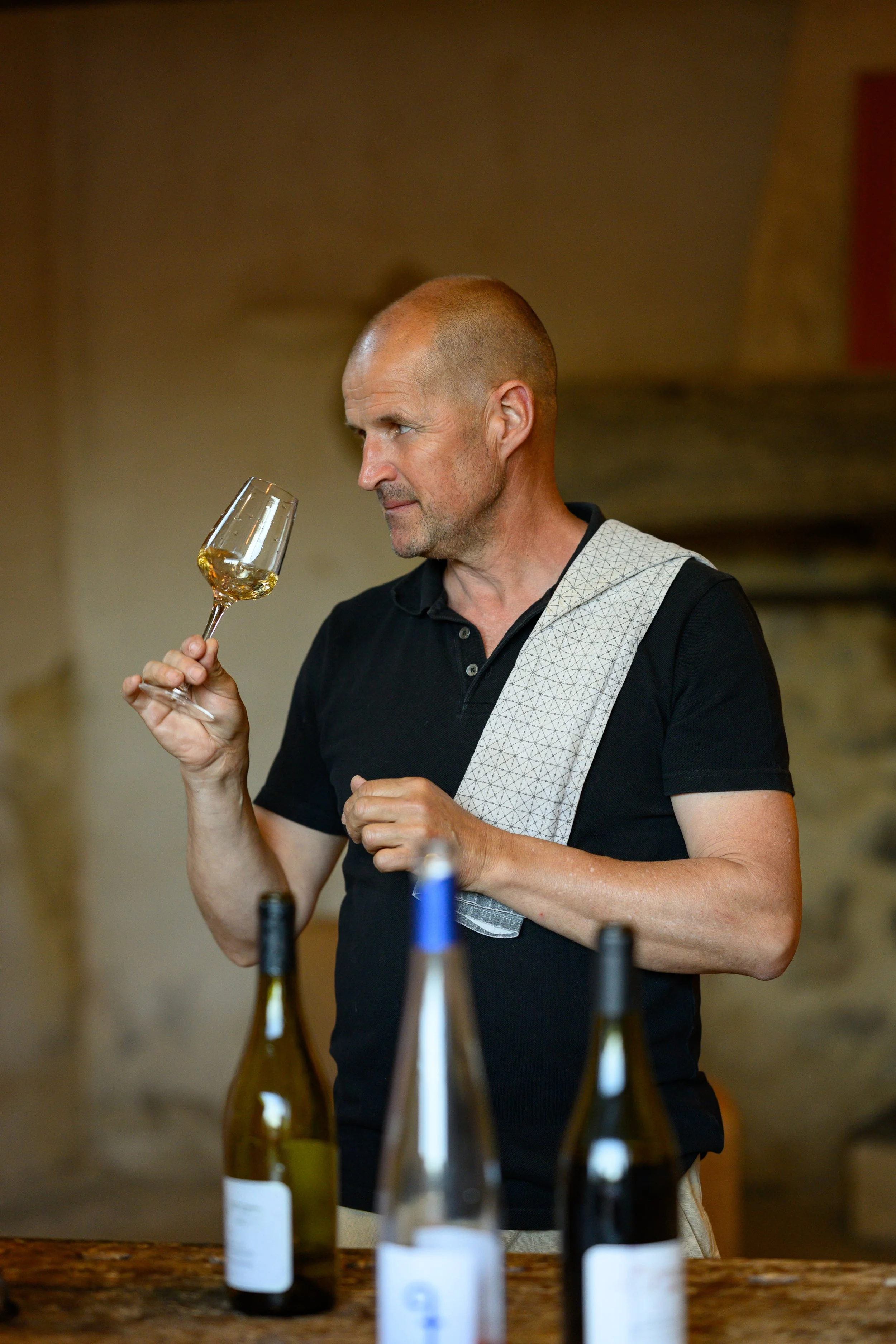A man with a shaved head and a short beard holding a glass of white wine, looking at it with a thoughtful expression. Several wine bottles are on the table in front of him.