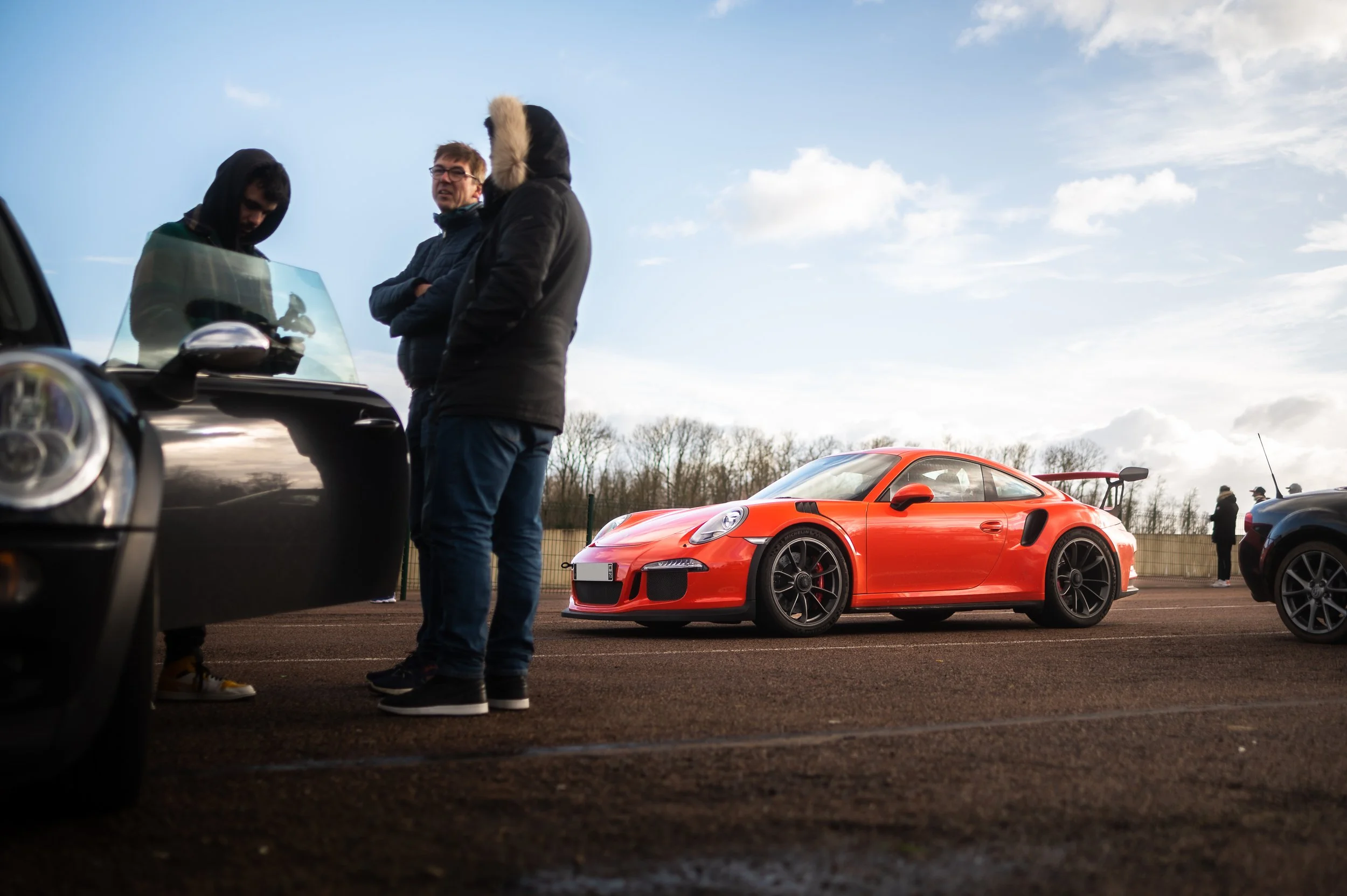 Three people standing next to a black car, with a red Porsche 911 sports car in the background on a parking lot under a partly cloudy sky.