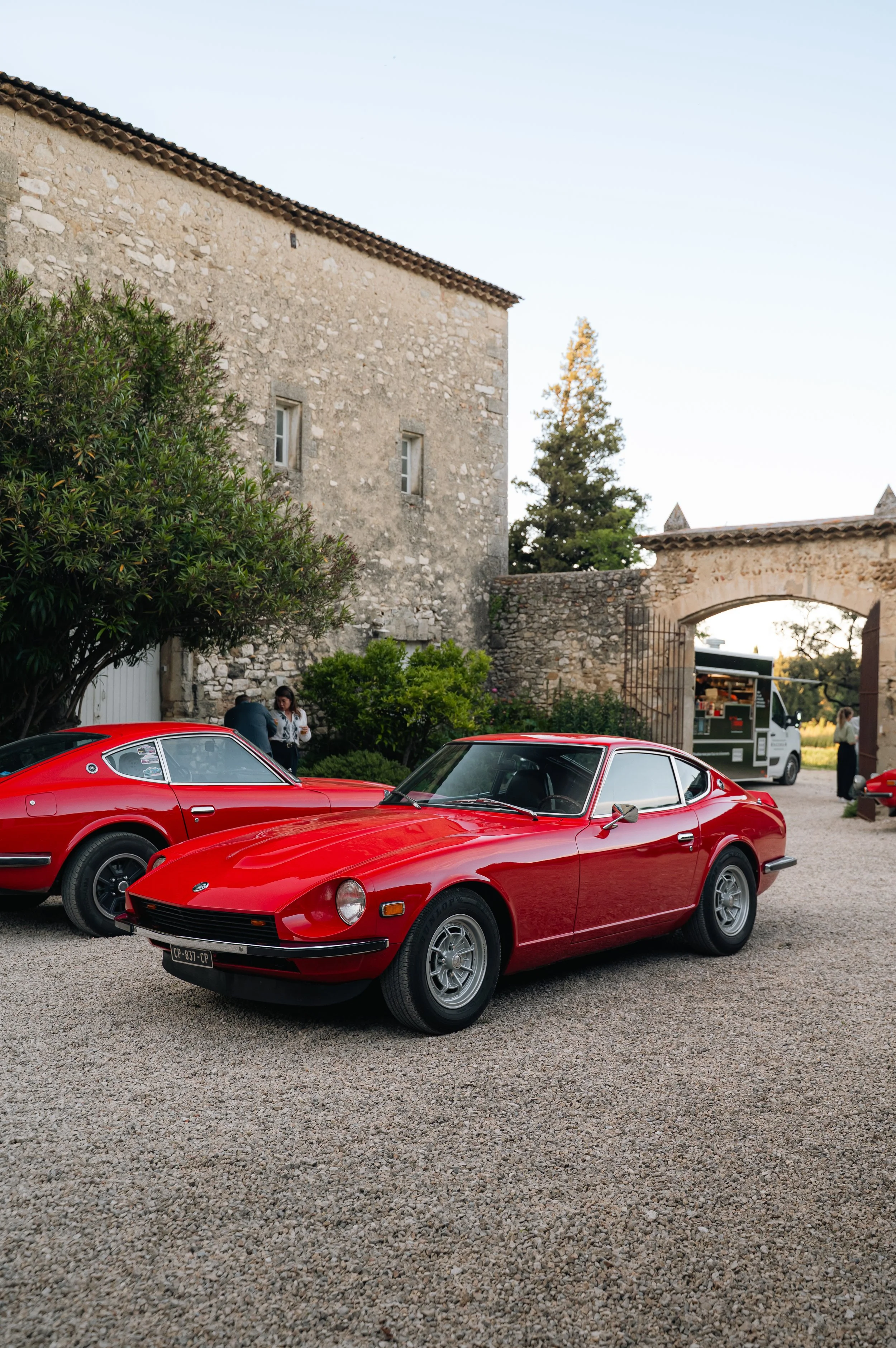 A red vintage sports car parked on a gravel surface in front of an old stone building with an archway, alongside another red vintage car, with a food truck and people in the background.