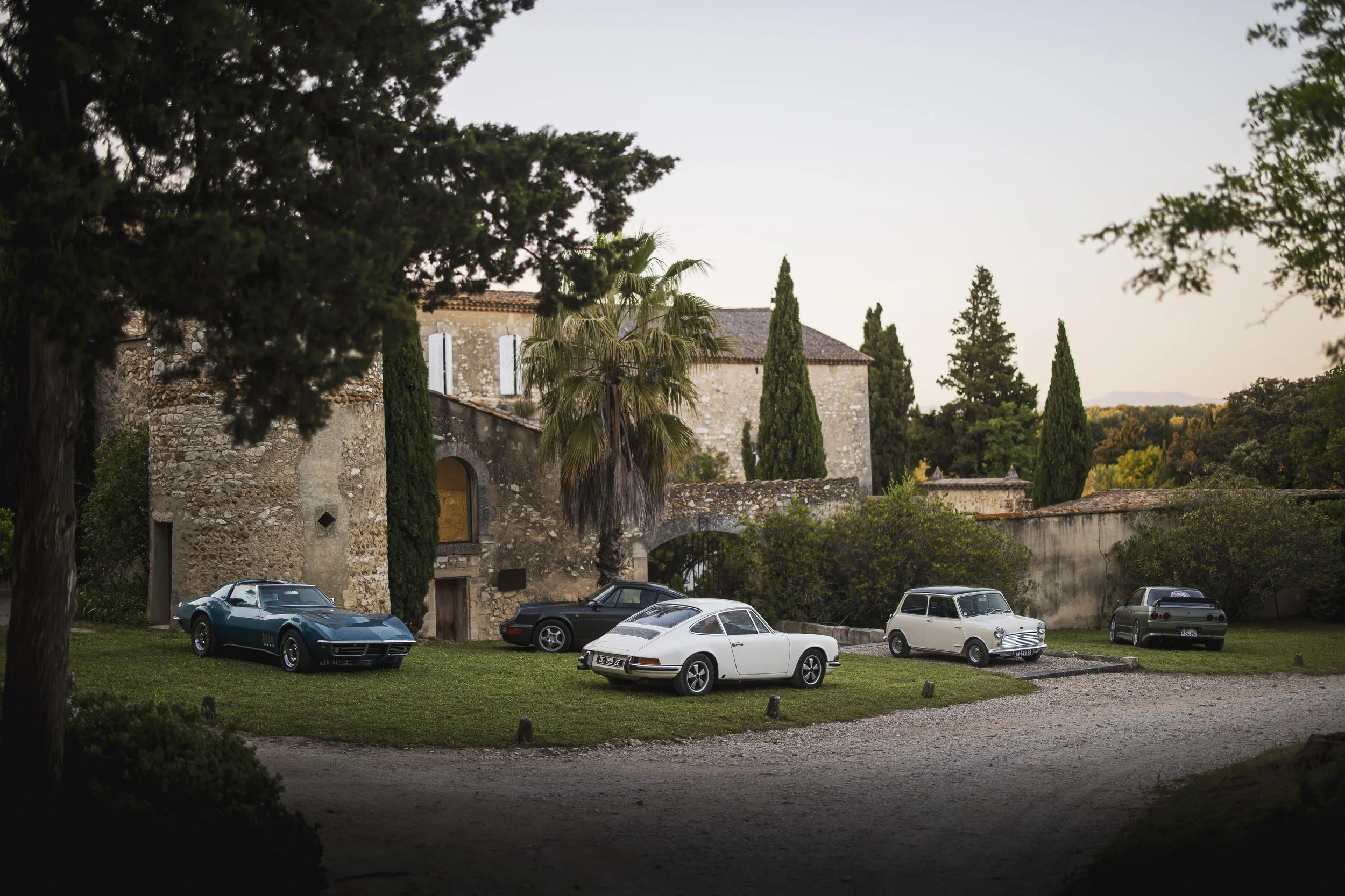 A collection of five vintage cars parked on a grassy area in front of an old stone building and a stone wall, surrounded by trees and foliage at dusk.