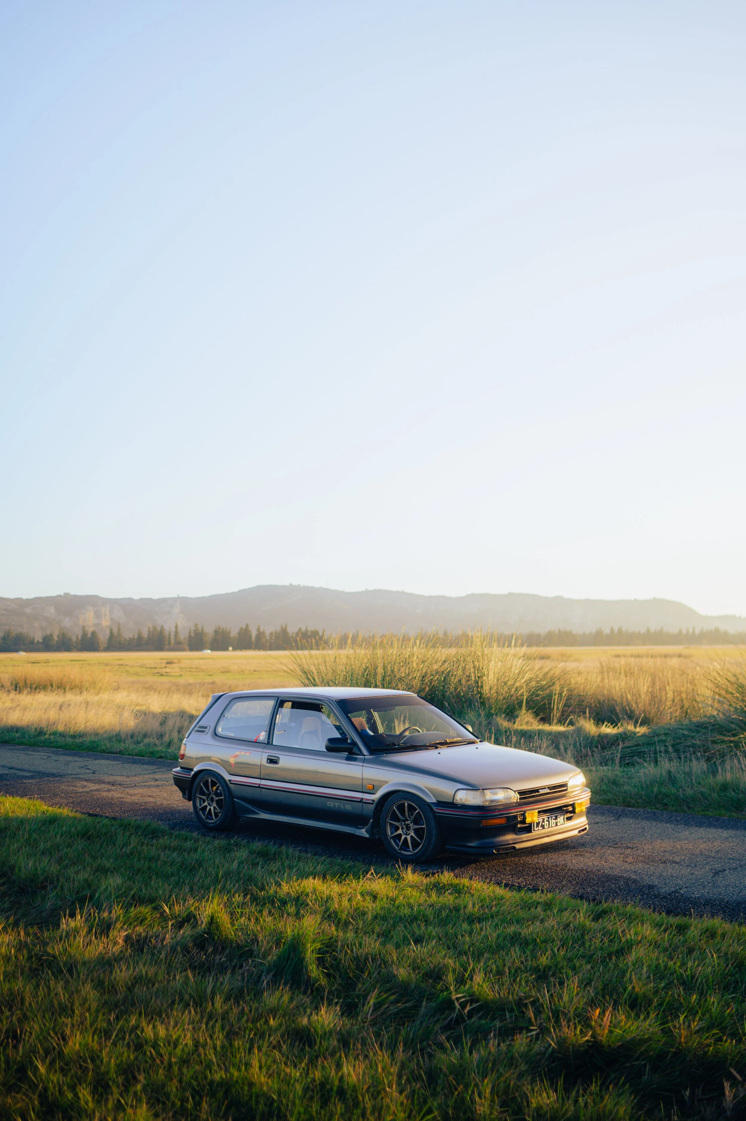 A Toyota Corolla GTI AE92 car parked on the side of a rural road with a vast field and distant mountains in the background, under a clear sky.