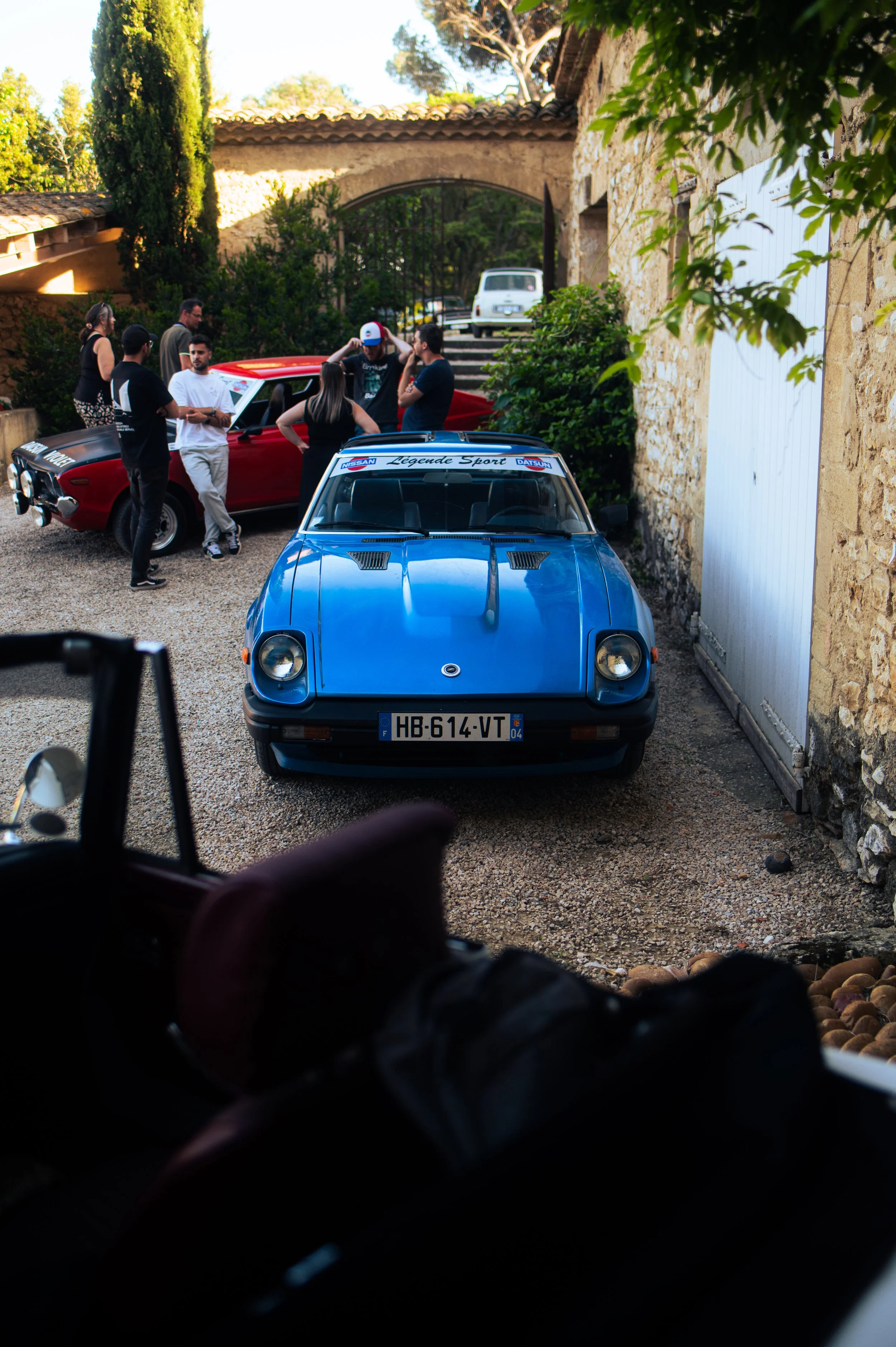 A group of people gather around a red Datsun 710SSS in a courtyard, with a blue Datsun 280ZX in the foreground and a stone building with a white door.
