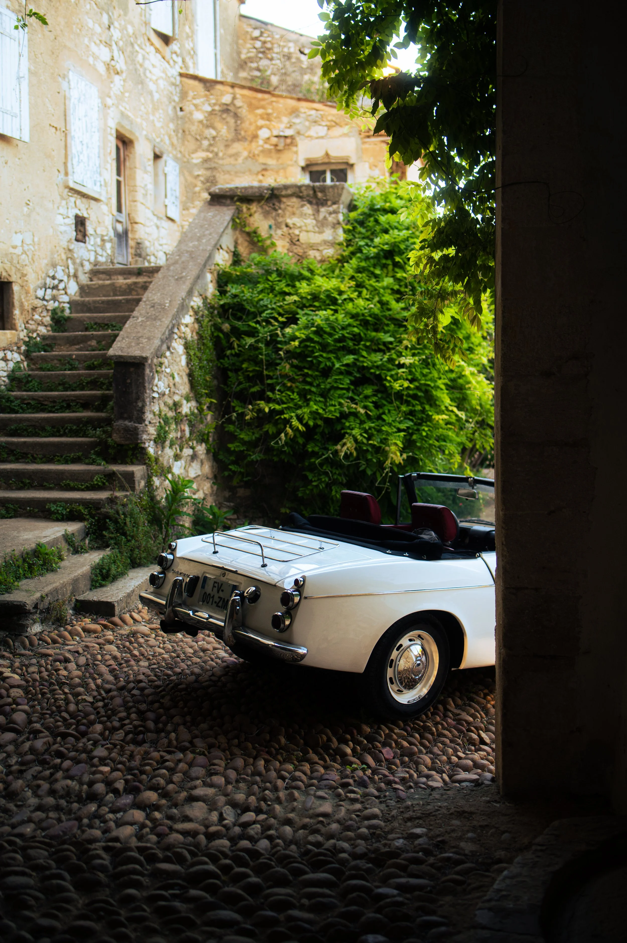 A white vintage Datsun Fairlady parked on a cobblestone street in a European village with stone buildings, stairs, and green foliage.