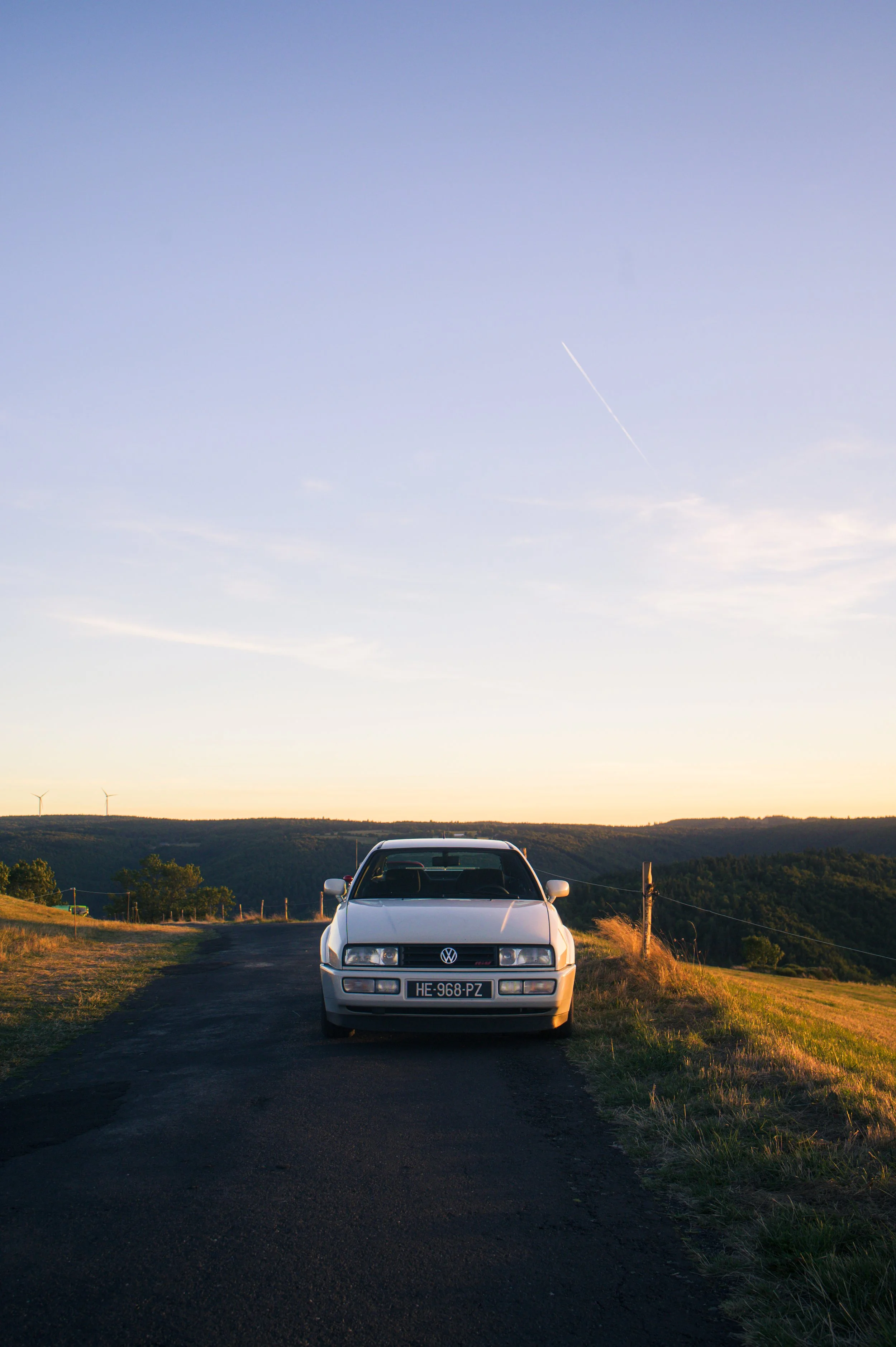 A white Volkswagen Corrado 16v car parked on a paved road with grassy sides, overlooking rolling hills and wind turbines in the distance at sunset.
