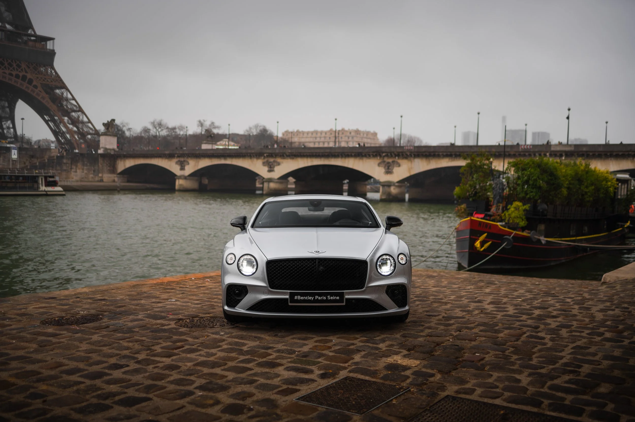 A silver Bentley Continental GT V8S parked on a cobblestone surface by the river, with the Eiffel Tower and a bridge in the background, overcast weather.