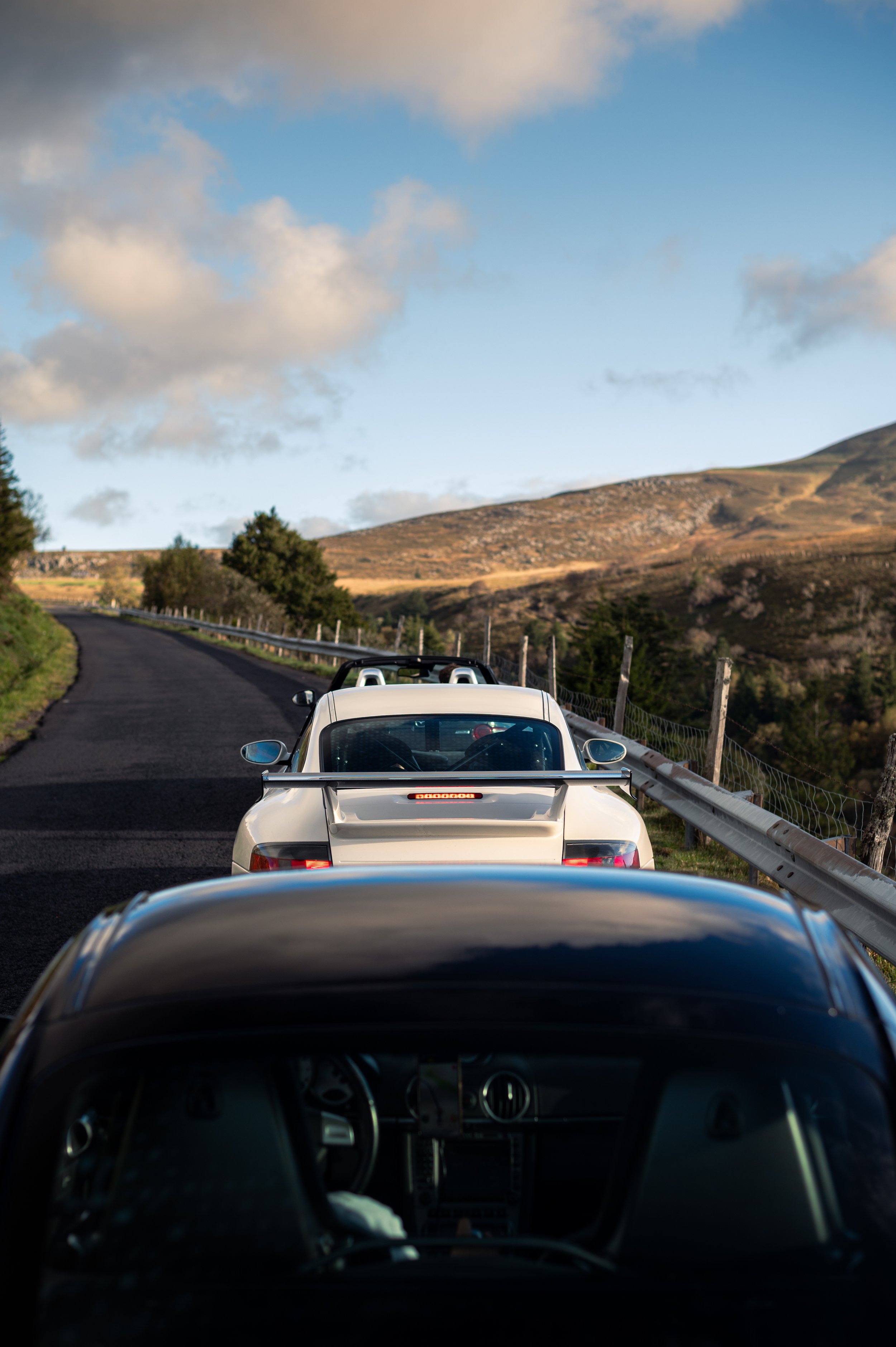 Two sports cars on a winding mountain road, with hills and trees in the background, under a partly cloudy sky.