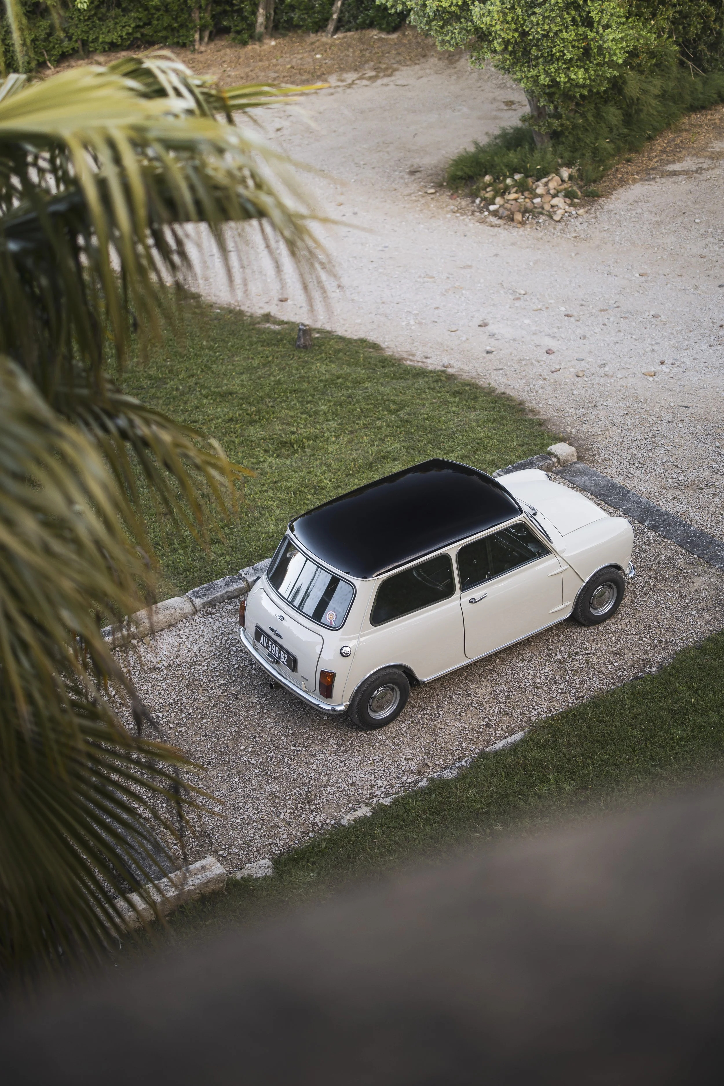 A vintage white car with a black roof parked on a gravel driveway, surrounded by greenery and trees.