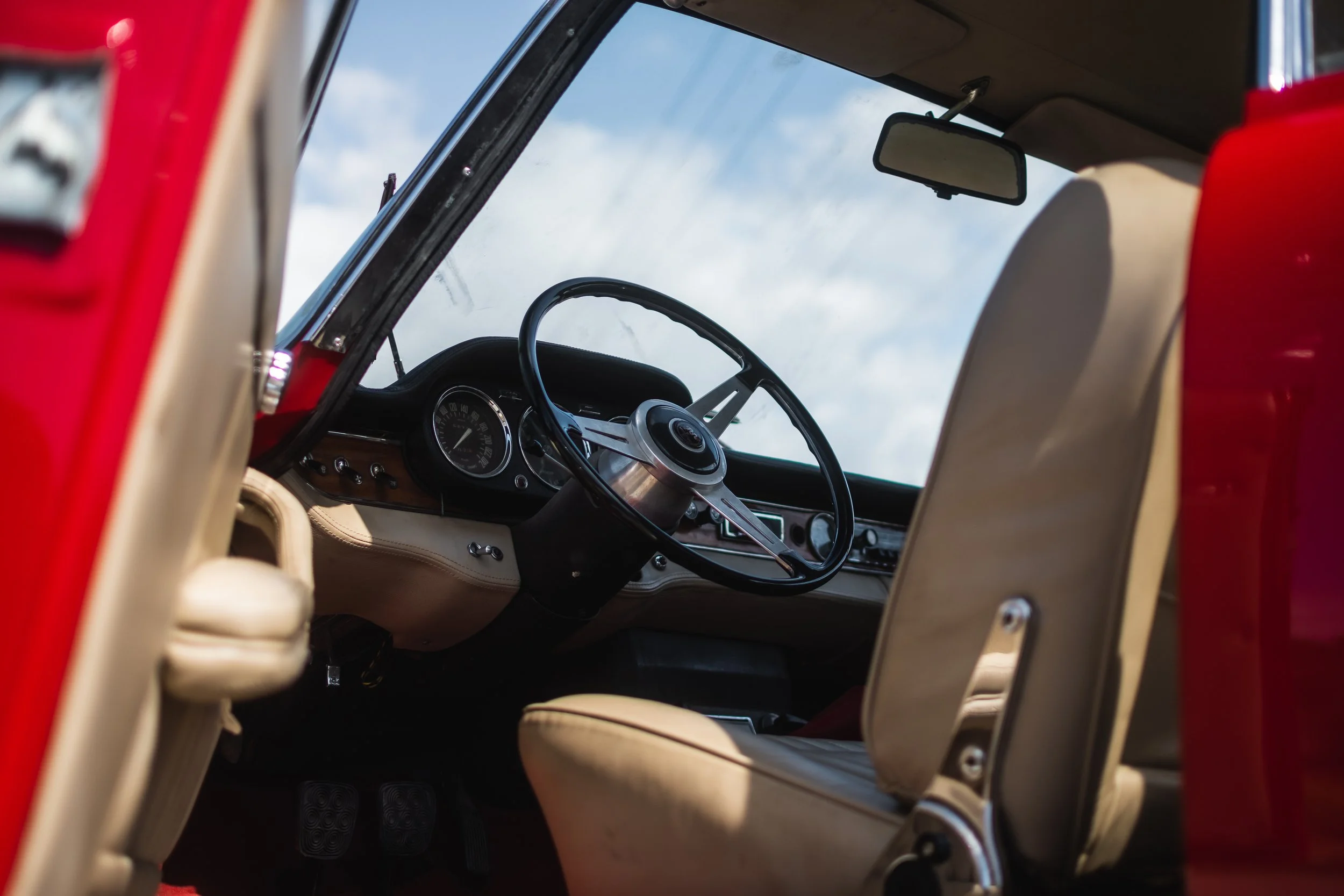 The interior of a Alfa Romeo 2600 Sprint showing the steering wheel, dashboard, and front seat with beige upholstery, viewed from the passenger side with the sky visible through the windshield.
