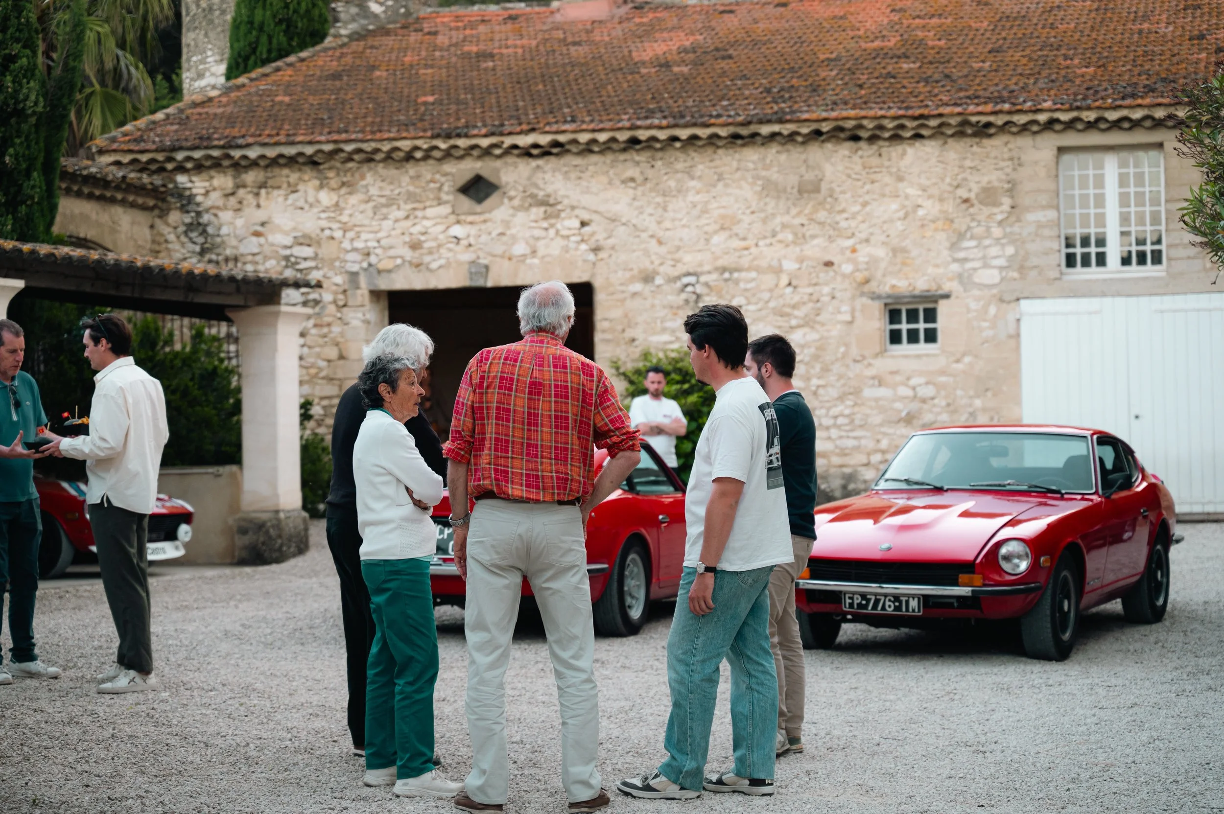 Group of people standing outside near vintage red cars, talking and socializing in front of a rustic stone building.