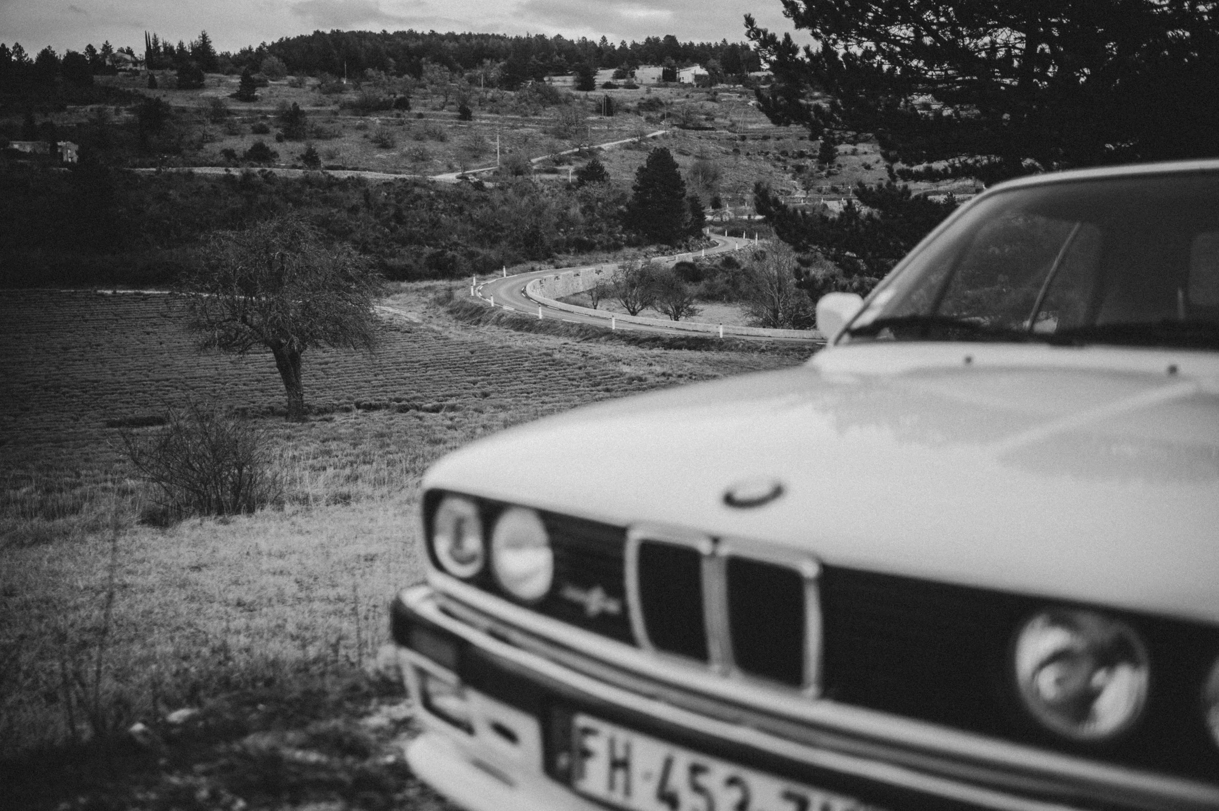 A black and white photo of a vintage BMW car in the foreground with a landscape of rolling hills, trees, and a winding road in the background.