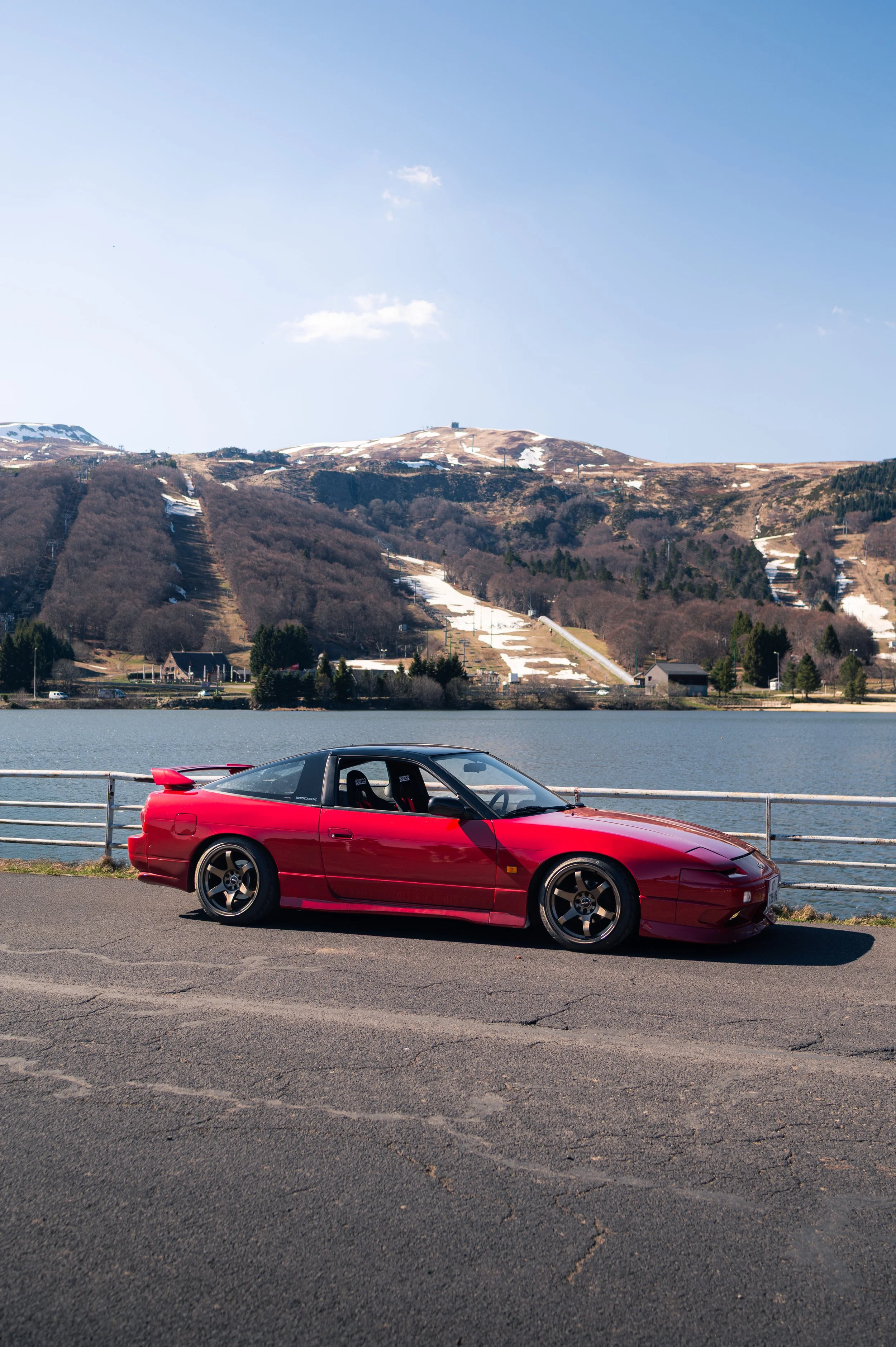 A red Nissan Silvia S13 parked beside a lake, with mountains and a partly cloudy sky in the background.