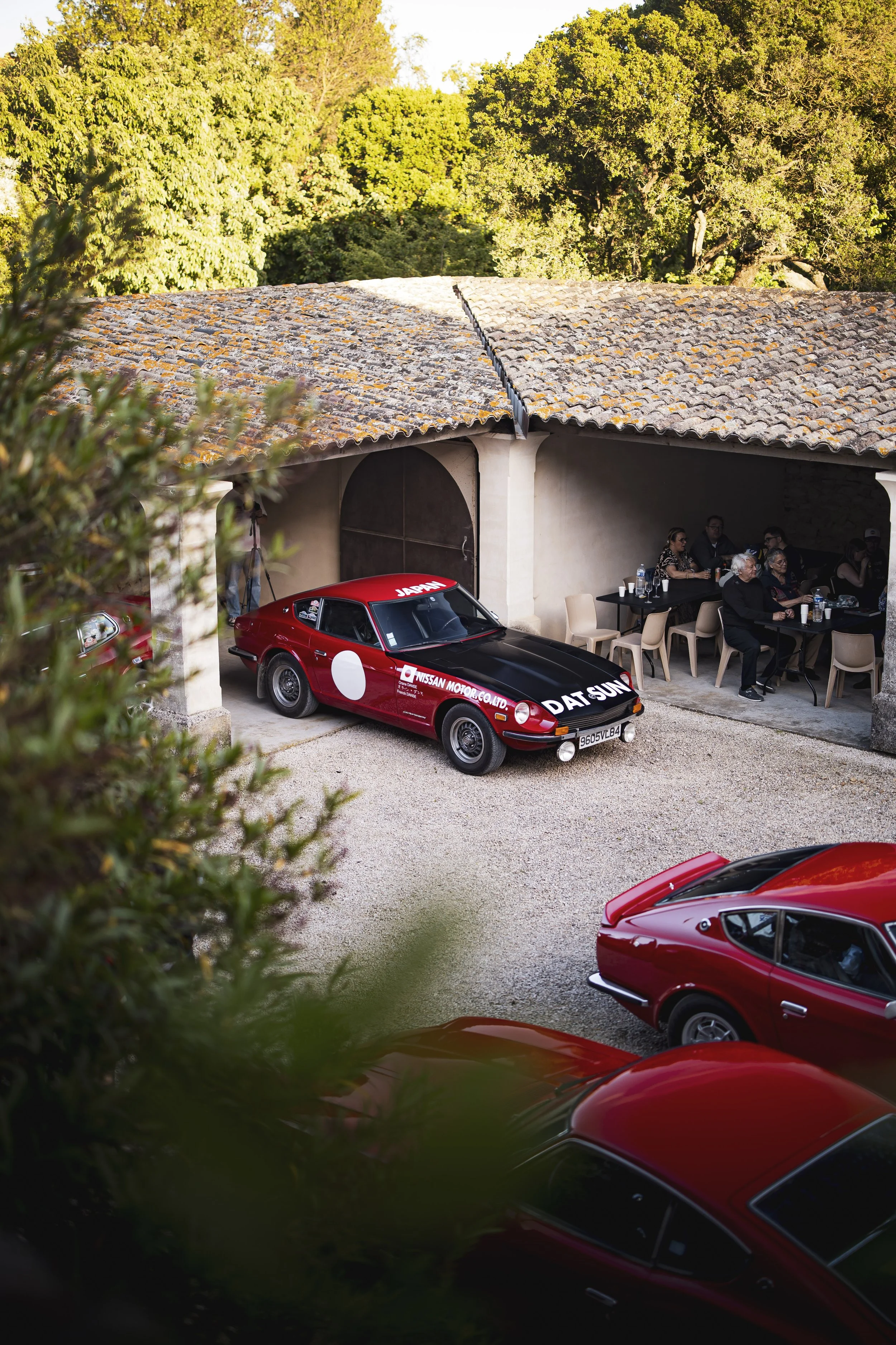 A red vintage race car with black accents parked partially in a garage, with people seated at a table in the background under a stone roof surrounded by greenery.