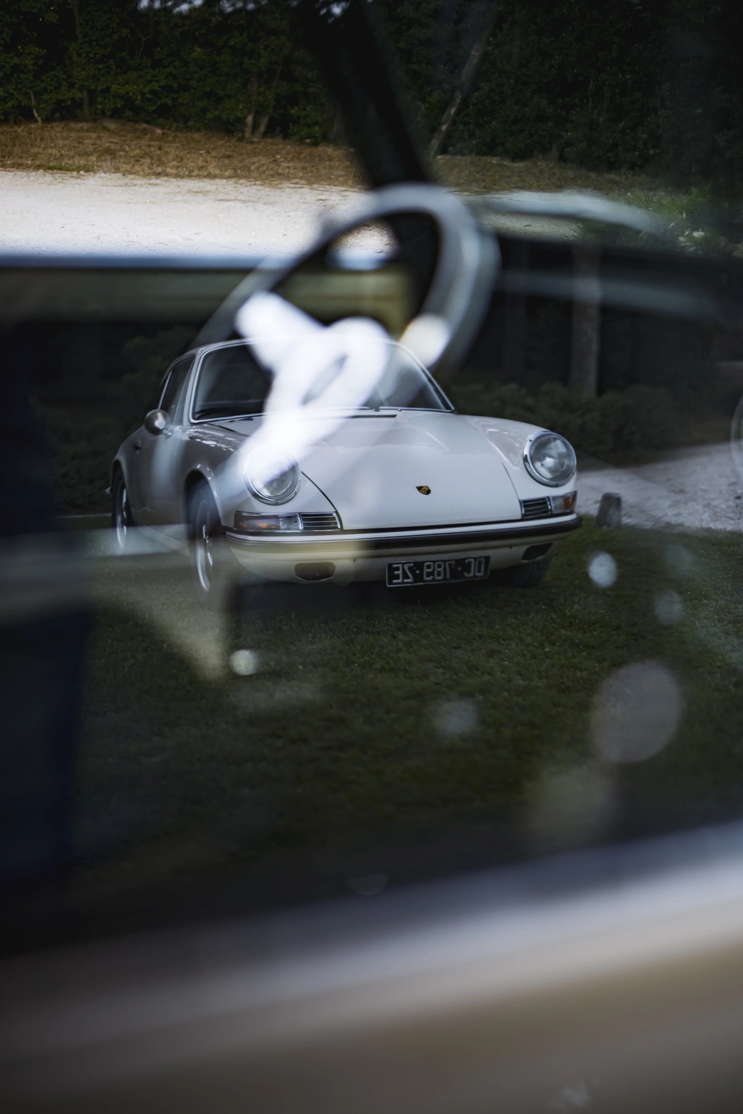 A classic white Porsche 911 viewed through the window of another vehicle, with the Porsche parked outside on a grassy area.