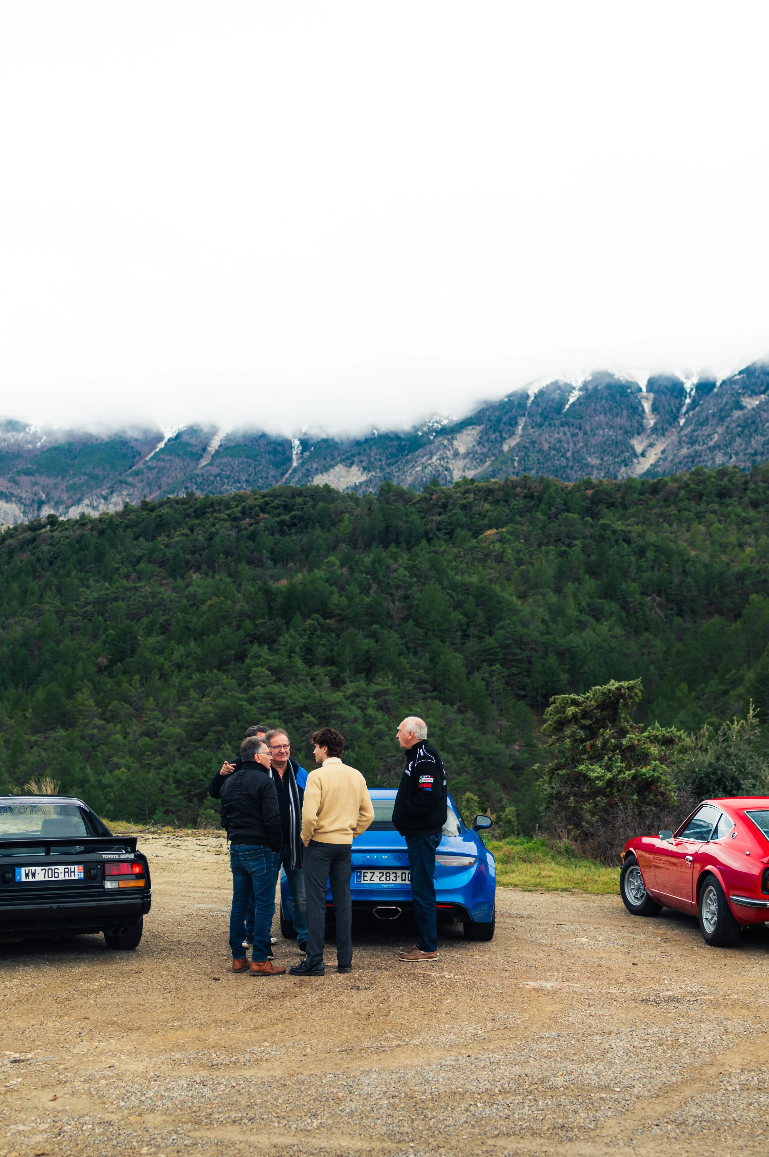 Group of five men standing and talking near parked sports cars on a dirt lot with green forest and mountain range in the background.