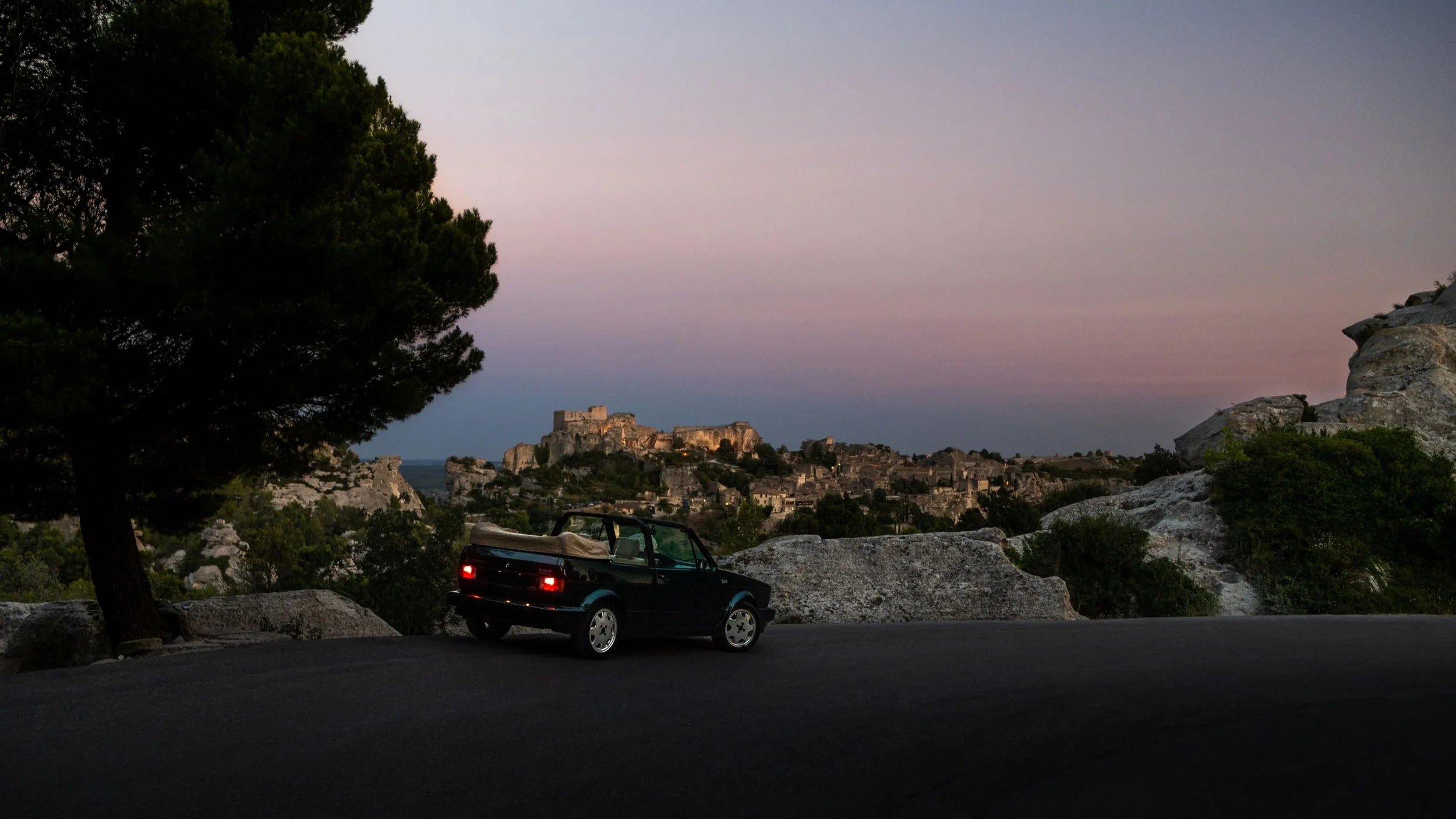 A small black convertible car parked on a scenic overlook with a castle on a hill in the distance during sunset or dusk.