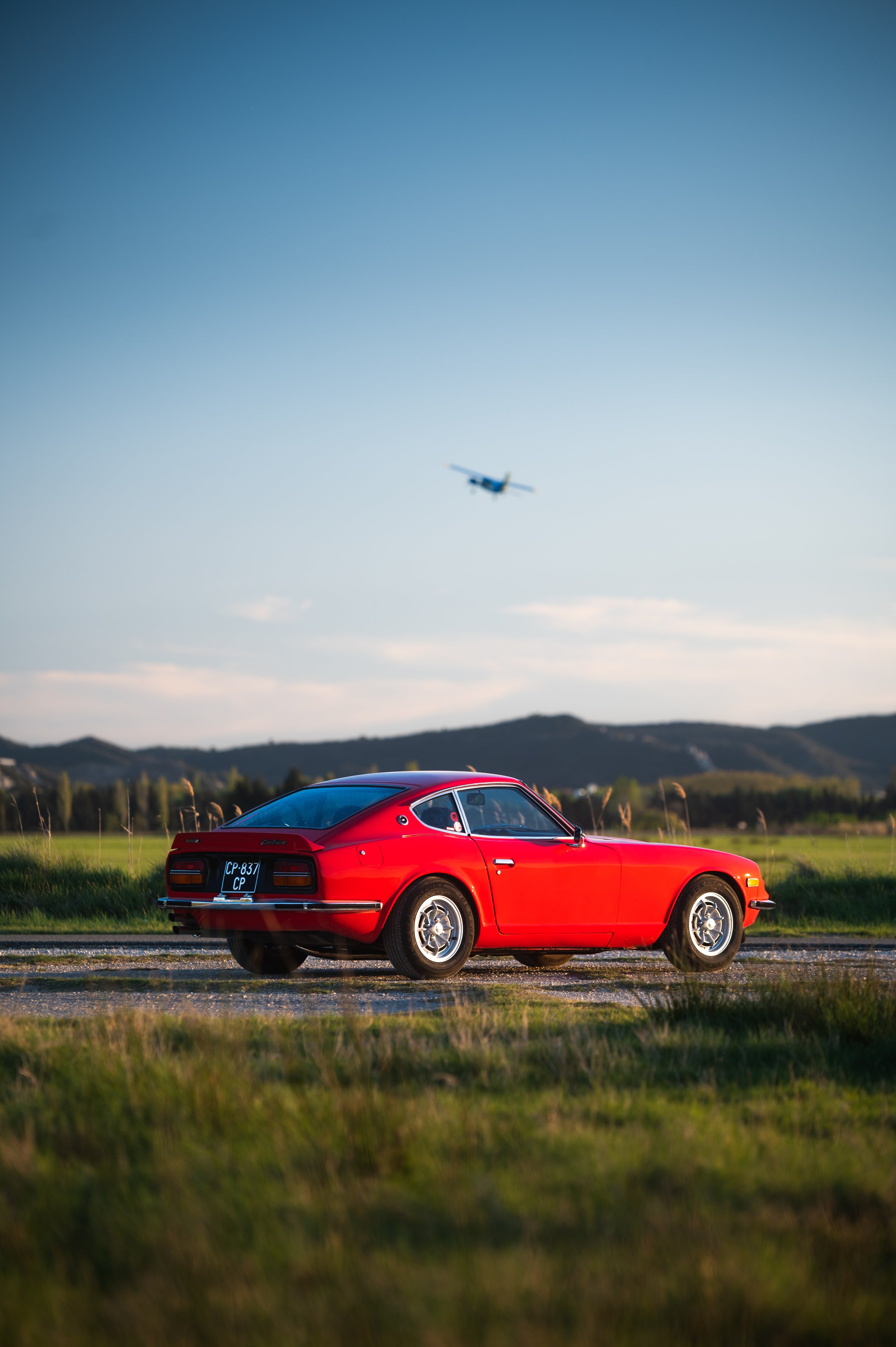 A red Datsun 240Z parked on a grassy field with mountains and an airplane flying in the sky above.