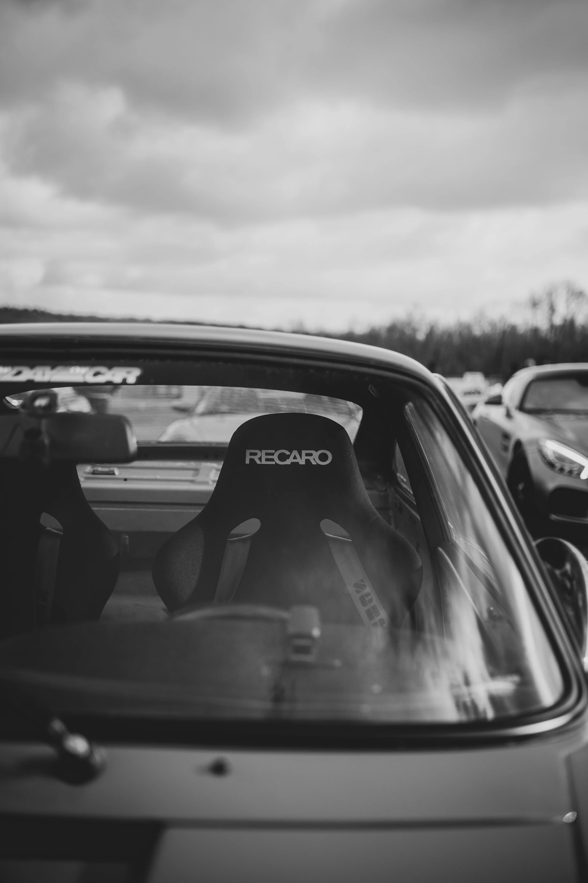 Black and white photo of the interior of a car, showing a Recaro racing seat through the windshield, with other cars and clouds in the background.