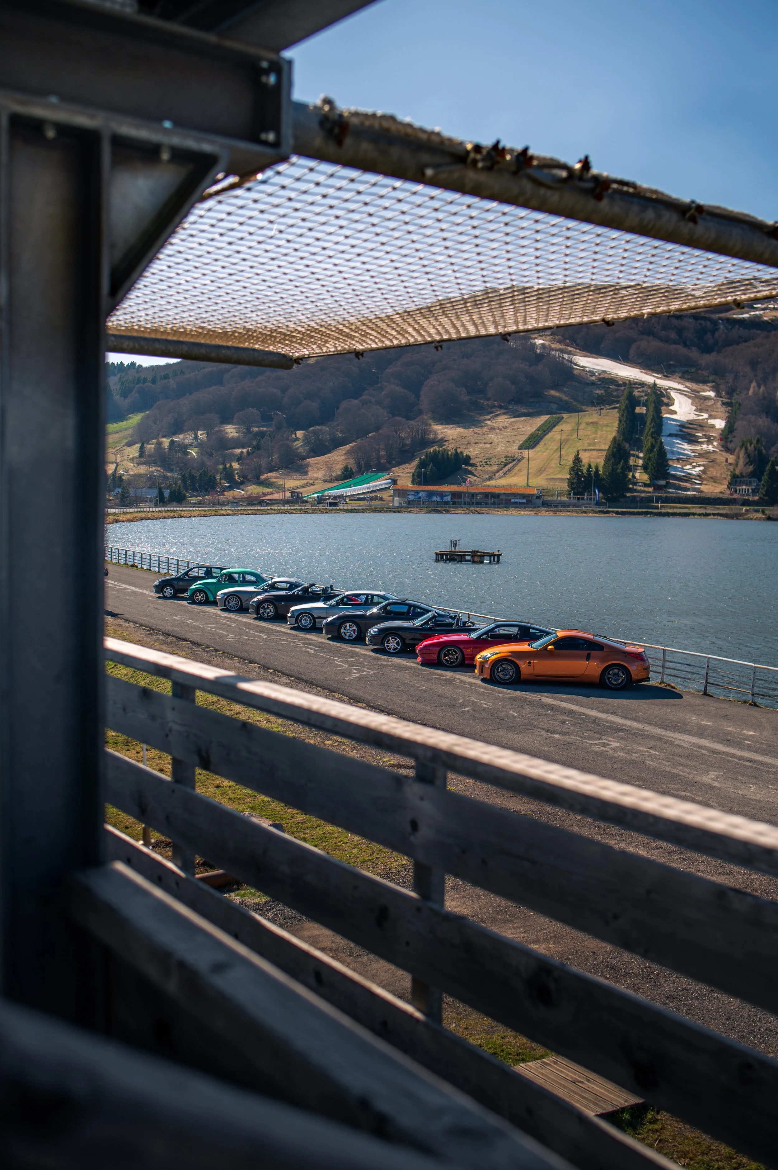 A line of sports cars parked along the edge of a body of water, viewed through the structure of a boat with a netted canopy, with hills and ski slopes in the background.