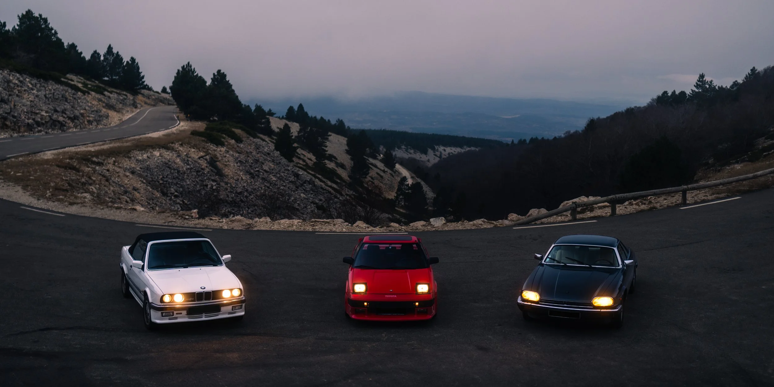 Three vintage cars parked on a mountain road at dusk with a scenic view of hills and trees in the background.