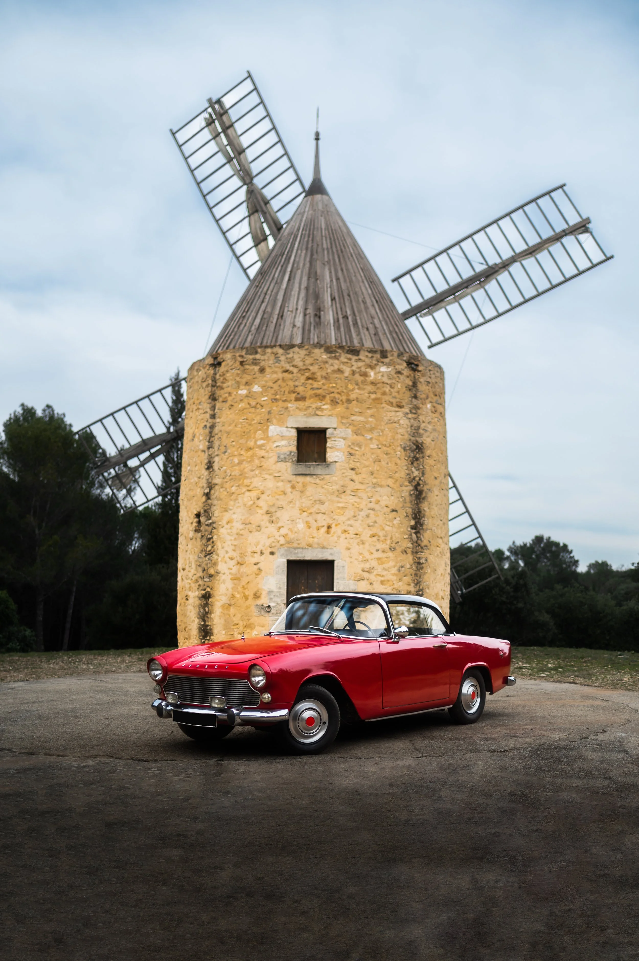 A red Simca Aronde parked in front of a small stone windmill with wooden blades.