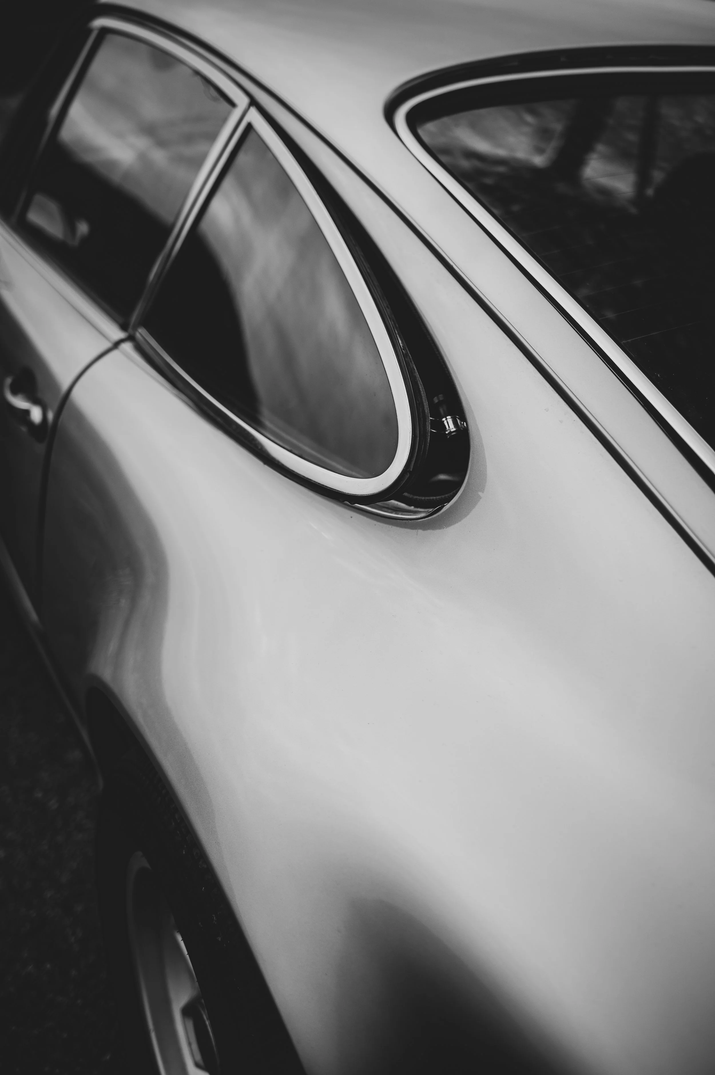 Close-up of a vintage car's rear side, black and white photograph showing the back wheel and part of the car body with rounded window and chrome details.
